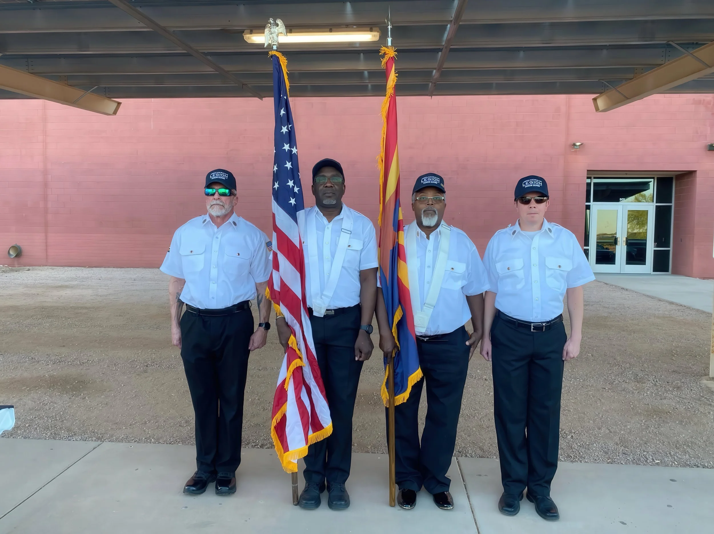 Four men standing in a row holding flags, with a brick building in the background. The men are dressed in white shirts and dark pants, with three wearing blue caps that read 'American Legion Honor Guard.' Two flags are displayed: the United States fl