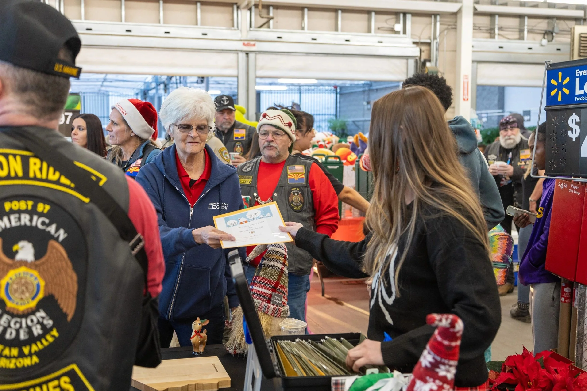 Group of people at a holiday event inside a store, some wearing Christmas hats, with a woman receiving a certificate from another woman, with festive decorations and gift items visible.