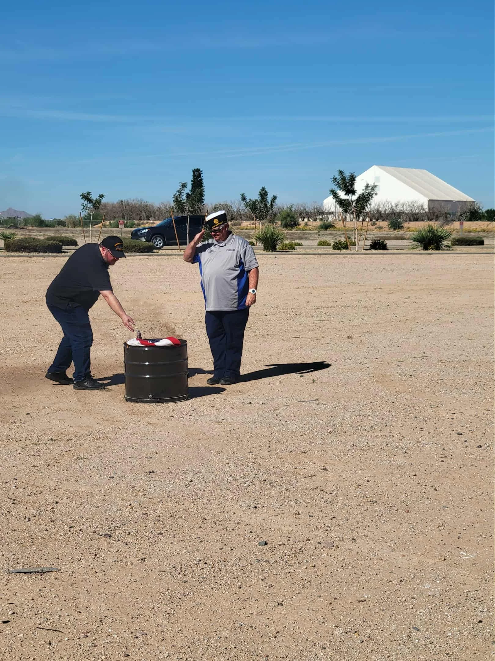 Two men in military uniforms setting up a flag on a flagpole in a dry, desert-like area with some plants and a warehouse in the background.
