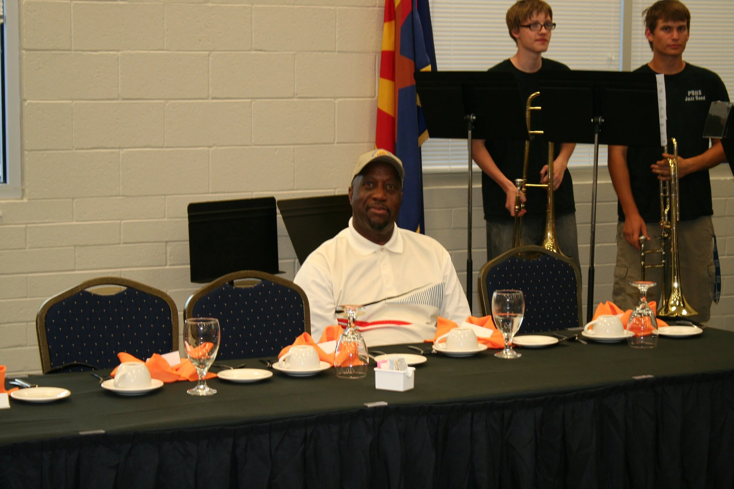 A man sitting at a banquet table with orange napkins, glasses, cups, and plates, with three men standing in the background holding musical instruments in front of a wall with an Arizona flag.