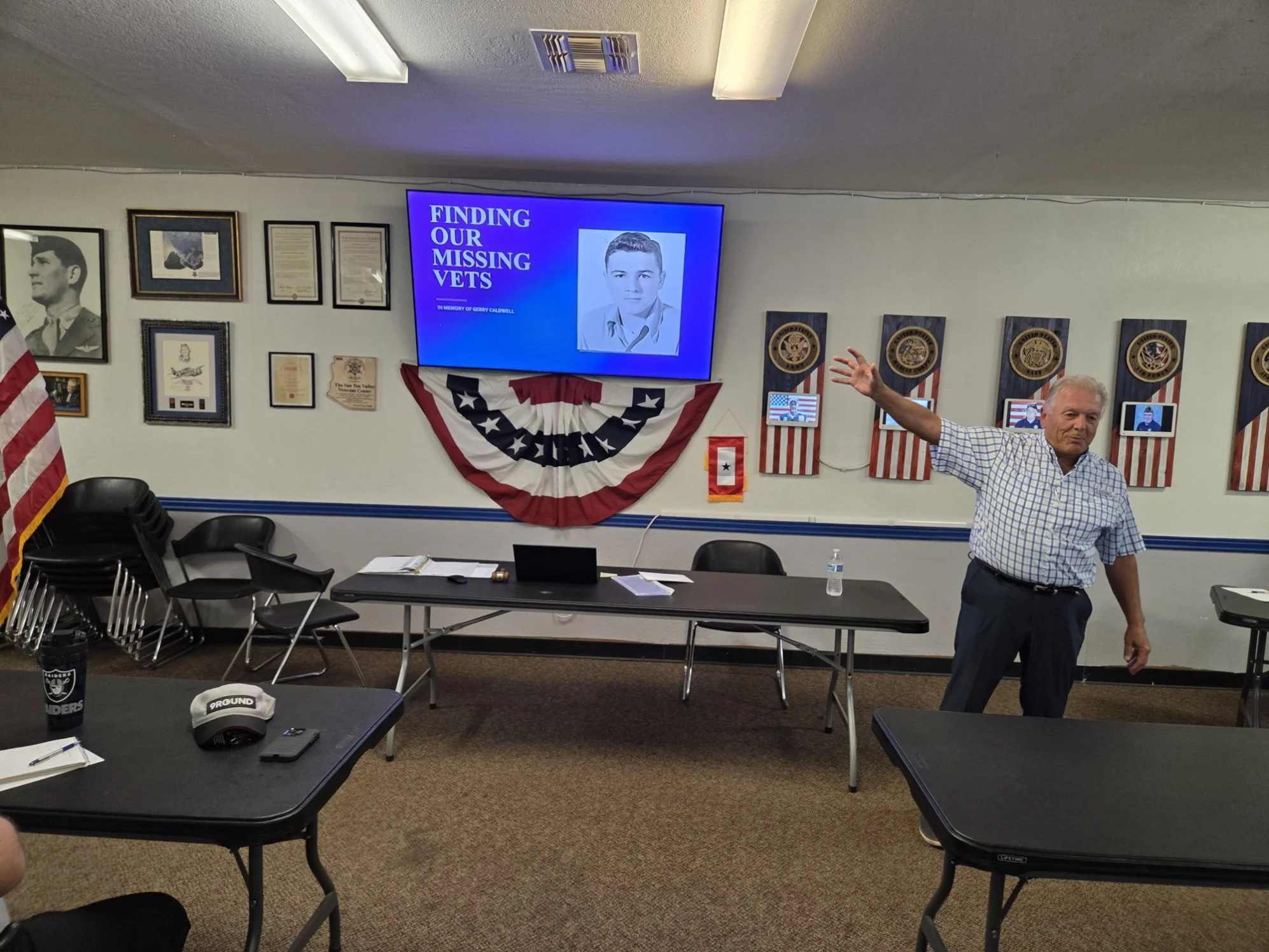 A room decorated for a patriotic event featuring a large screen with the text 'Finding Our Missing Vets' and a photo of a young man. The room has American flags, framed documents, and military emblems on the wall. An older man in a checkered shirt is