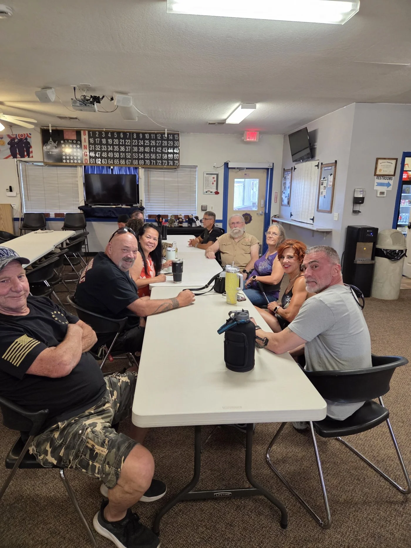A group of people sitting around a long rectangular table in a community room, smiling at the camera. The room has a bingo board, a large TV, and various signs on the wall.