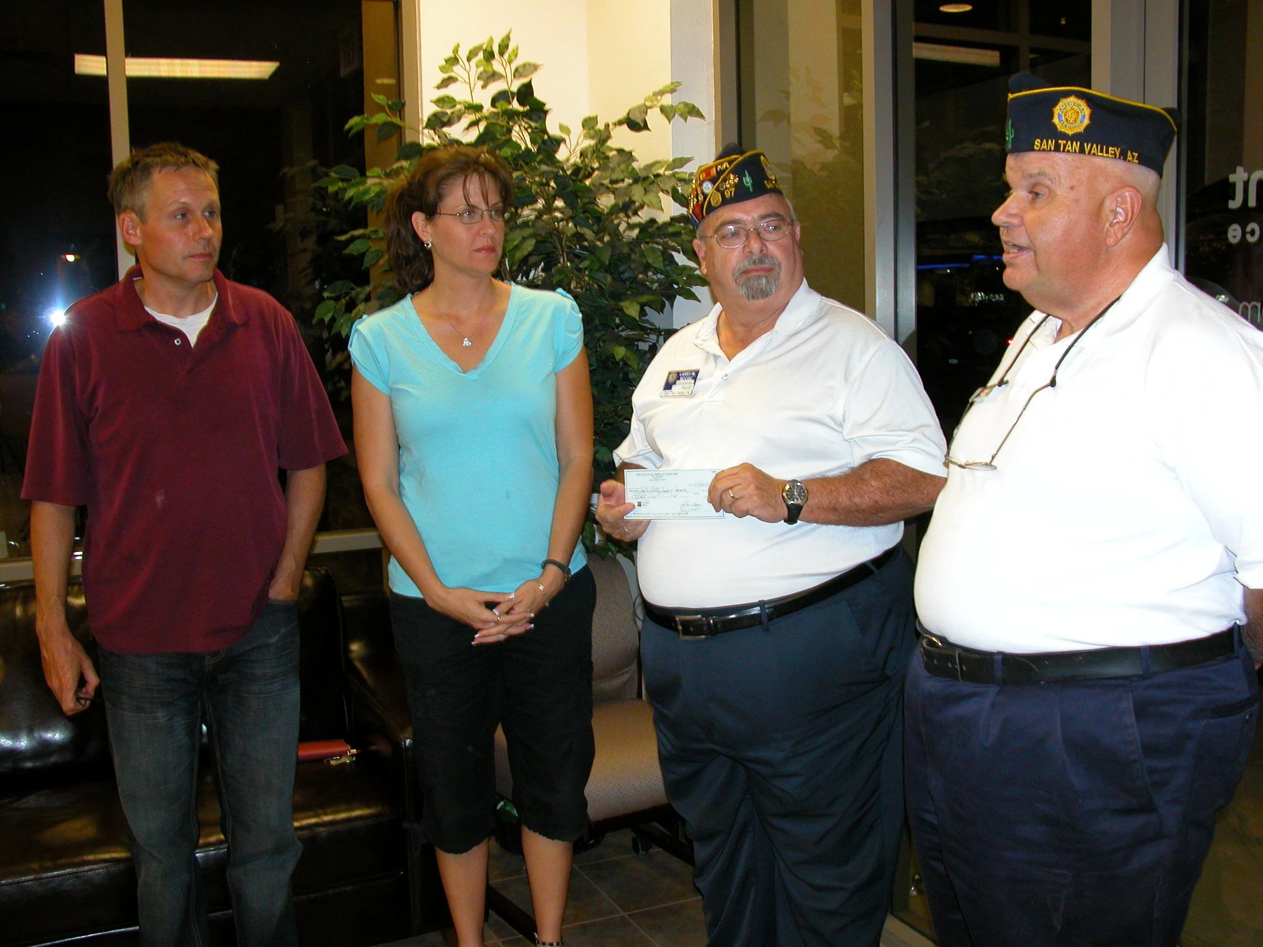 Four individuals engaged in conversation inside a building. The group includes two men wearing caps with insignia, one with a badge on his shirt, and a woman with glasses. They are standing in front of a plant and a window.