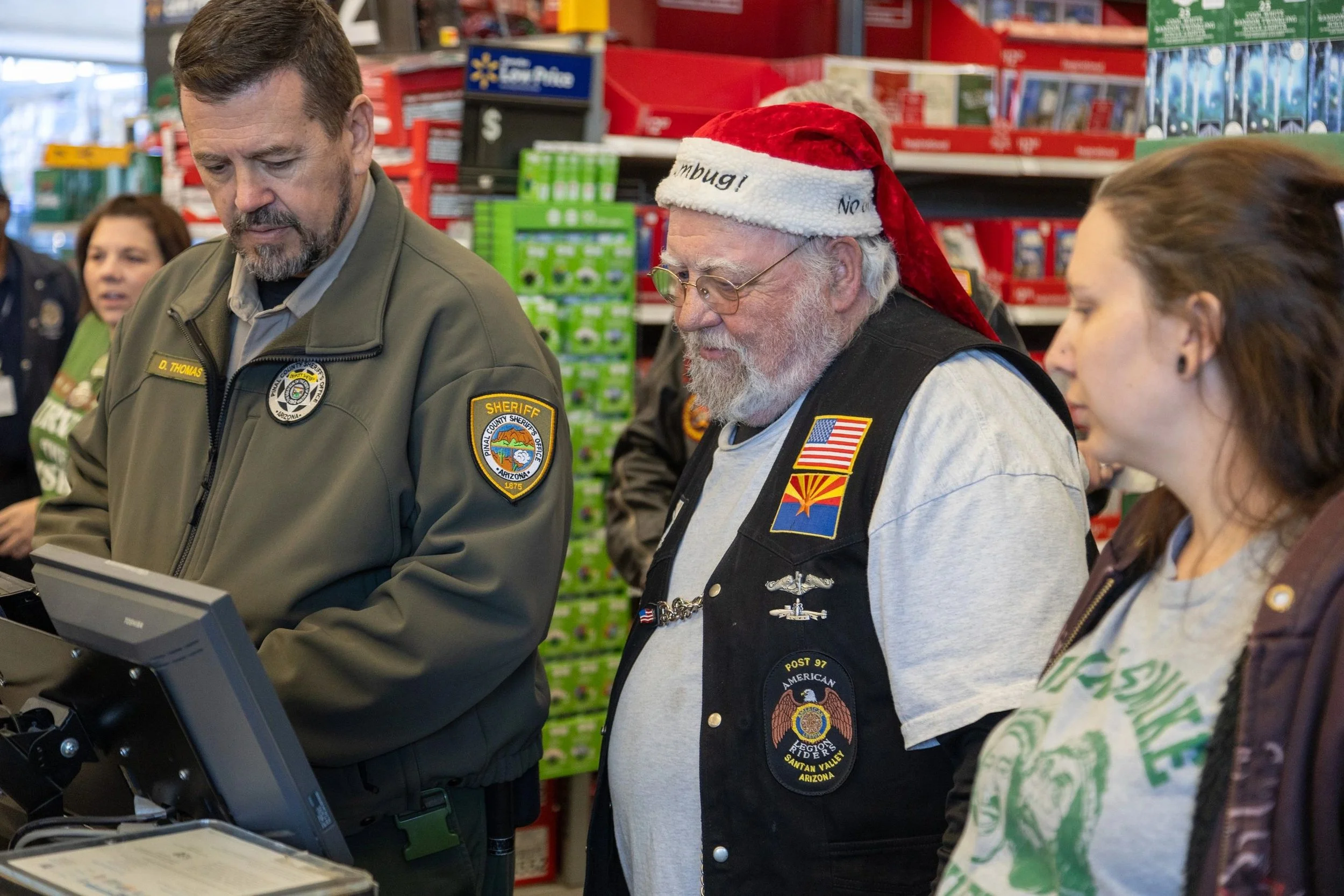 A group of people at a checkout counter in a store, including a man in a sheriff's uniform, a man wearing a Santa hat, and two women nearby.