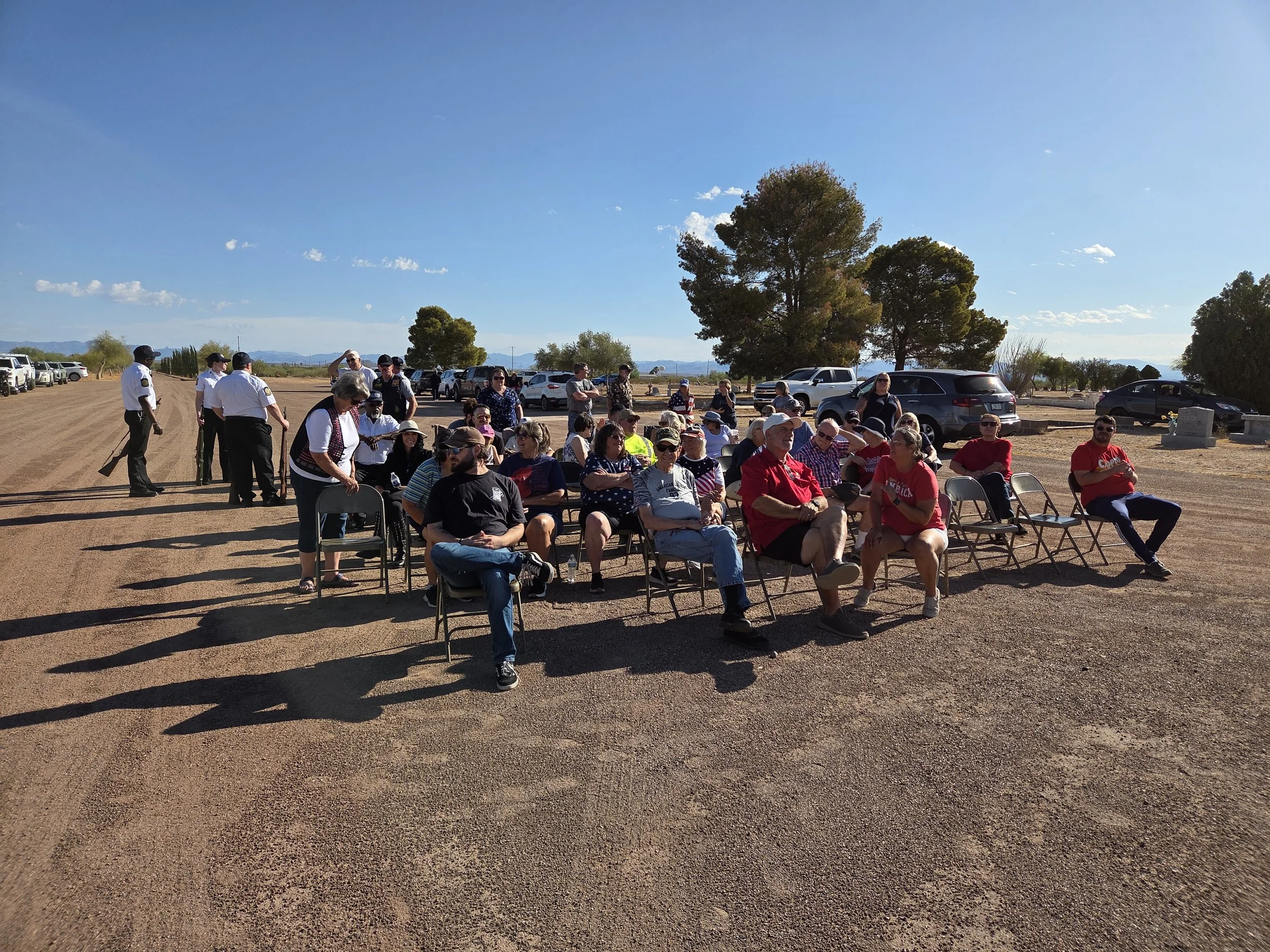 A group of people seated outdoors on folding chairs, gathered in a sunny open area with dirt ground, cars parked in the background, and large trees under a clear blue sky.