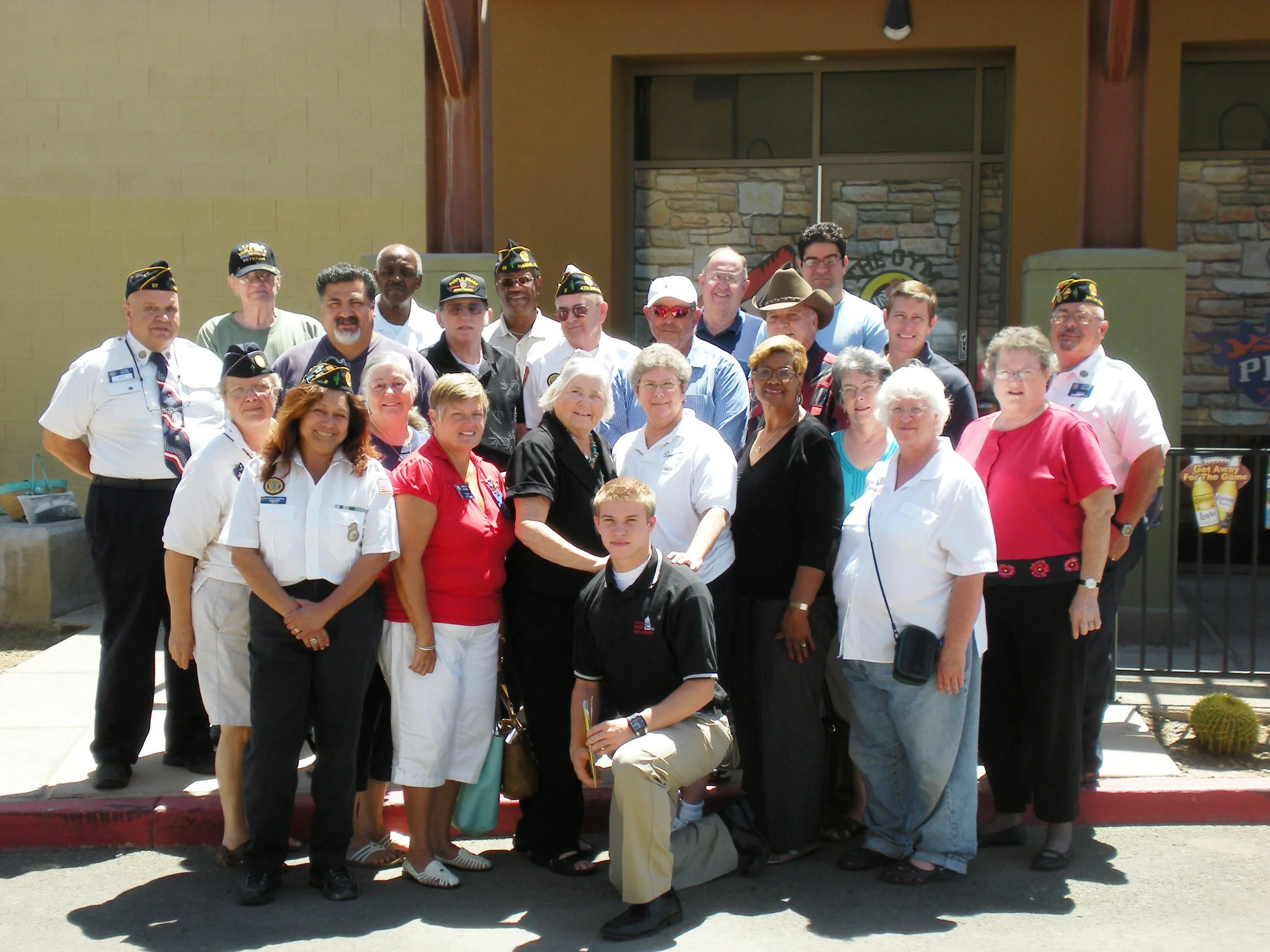 A group of people, some wearing veterans' hats, gathered outside a building with a stone and brick wall, posing for a photo.