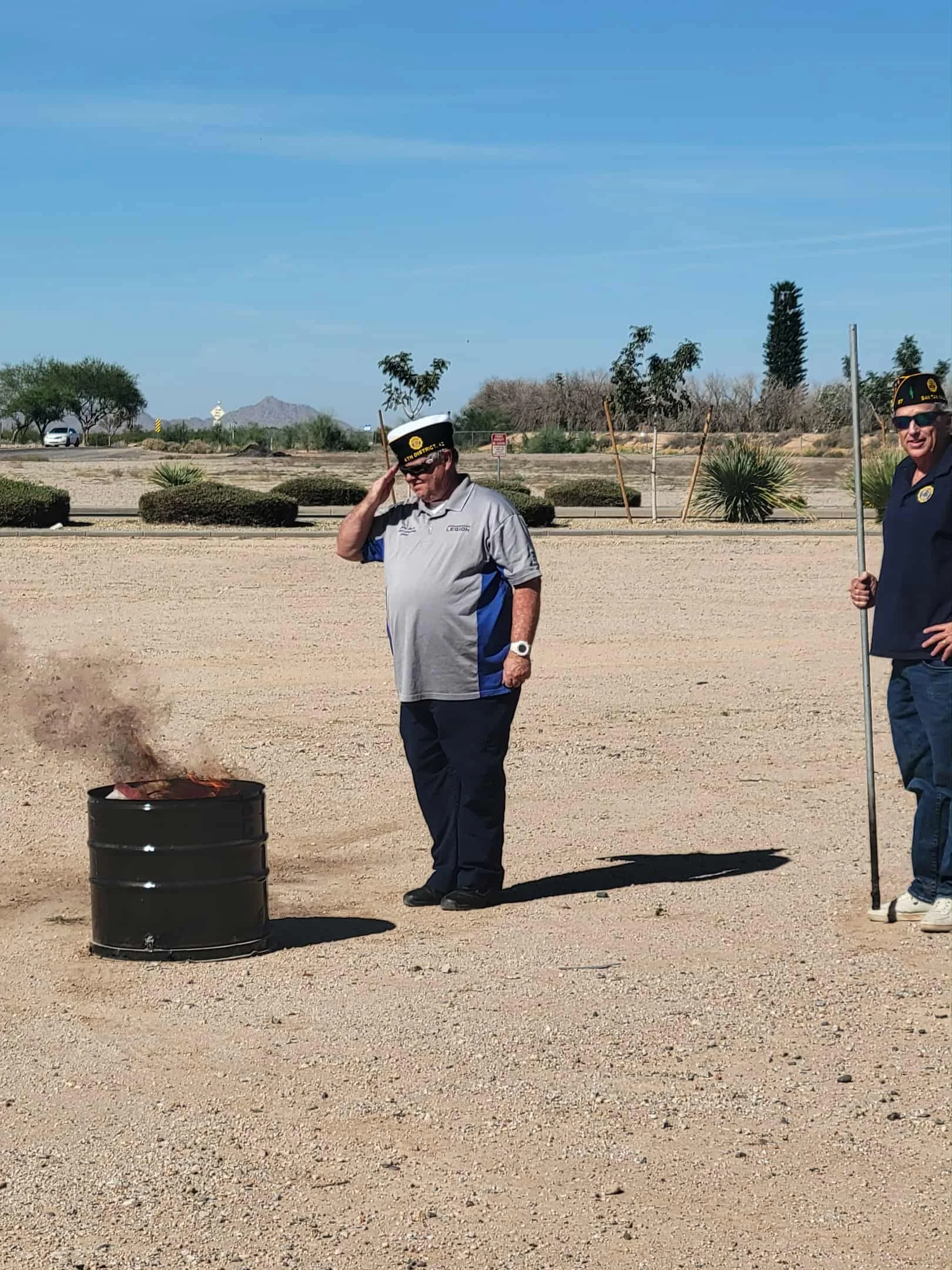 Two men wearing American Legion hats standing outdoors in a desert area with bushes, trees, and mountains in the background. One man salutes while the other stands with a flag pole. There is a black barrel in front of them.