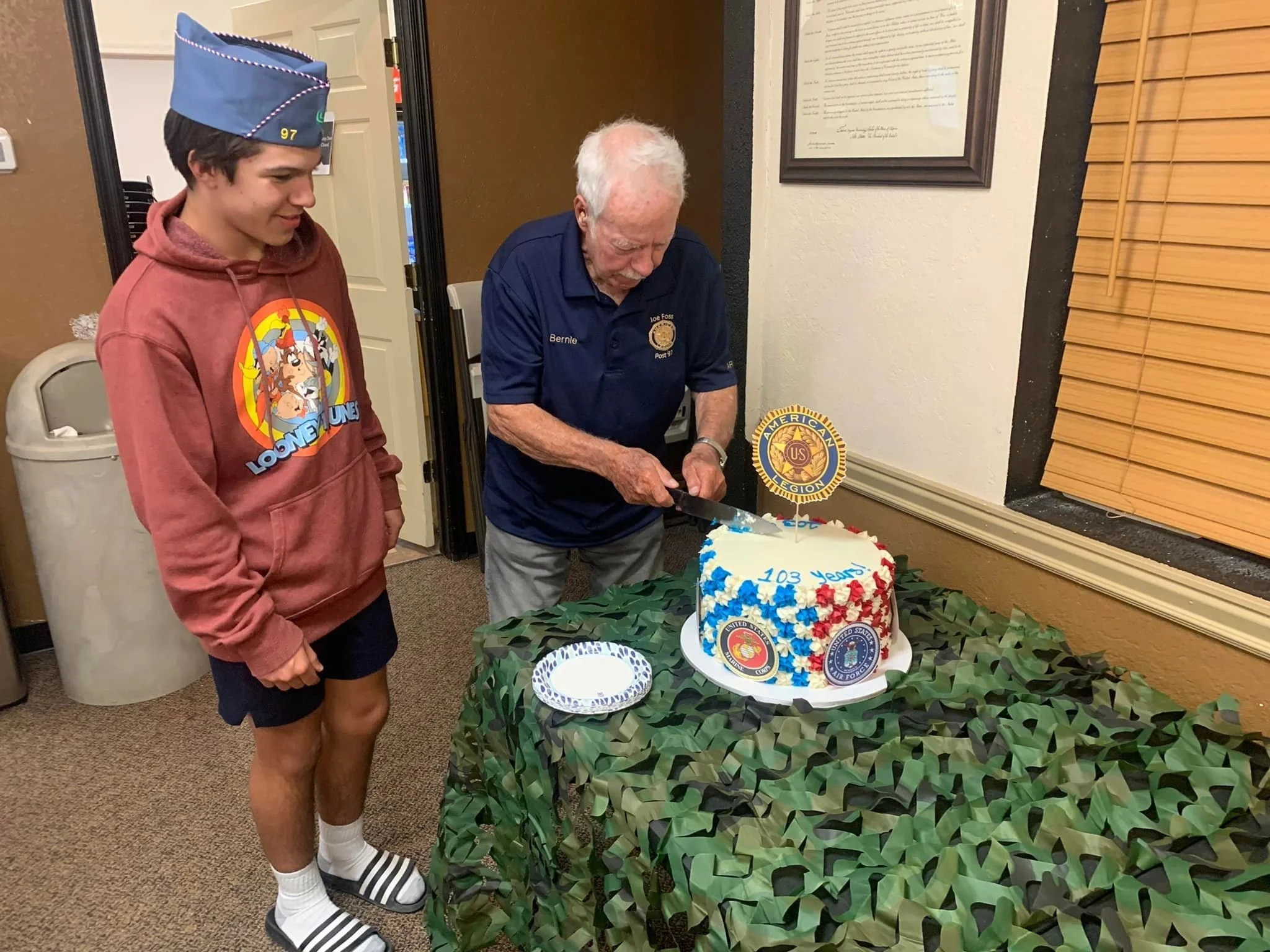 An elderly man with white hair wearing a navy blue shirt and a young man with brown hair wearing a pink hoodie with Looney Tunes graphic and blue graduation cap, stand close to a decorated table with a white cake that has blue, red, and white decorat