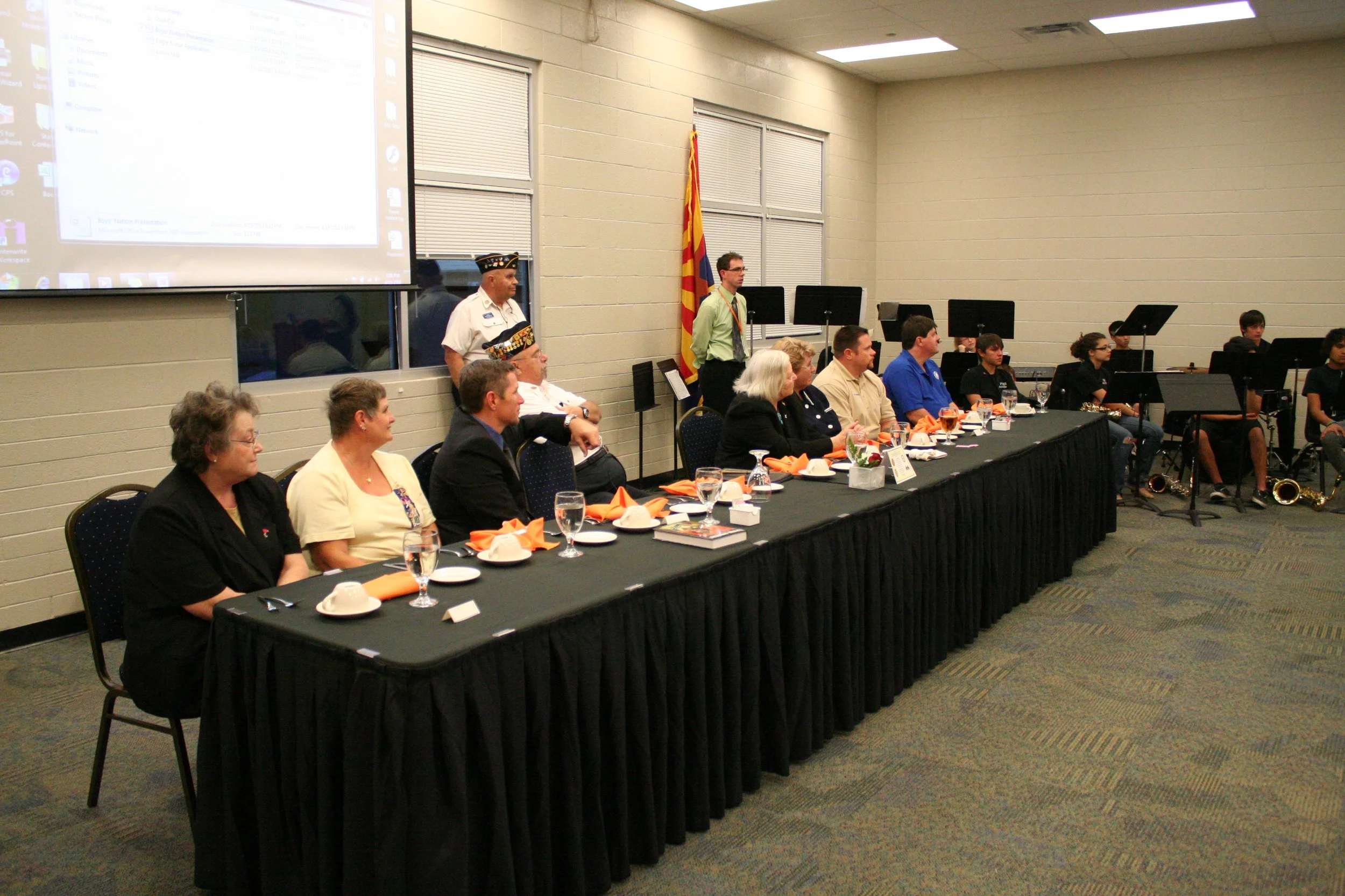 A large conference room with a long table set for a formal dinner, with glasses, plates, and orange napkins. People are seated along one side, some wearing business attire and military uniforms, facing a group of musicians with instruments at the bac