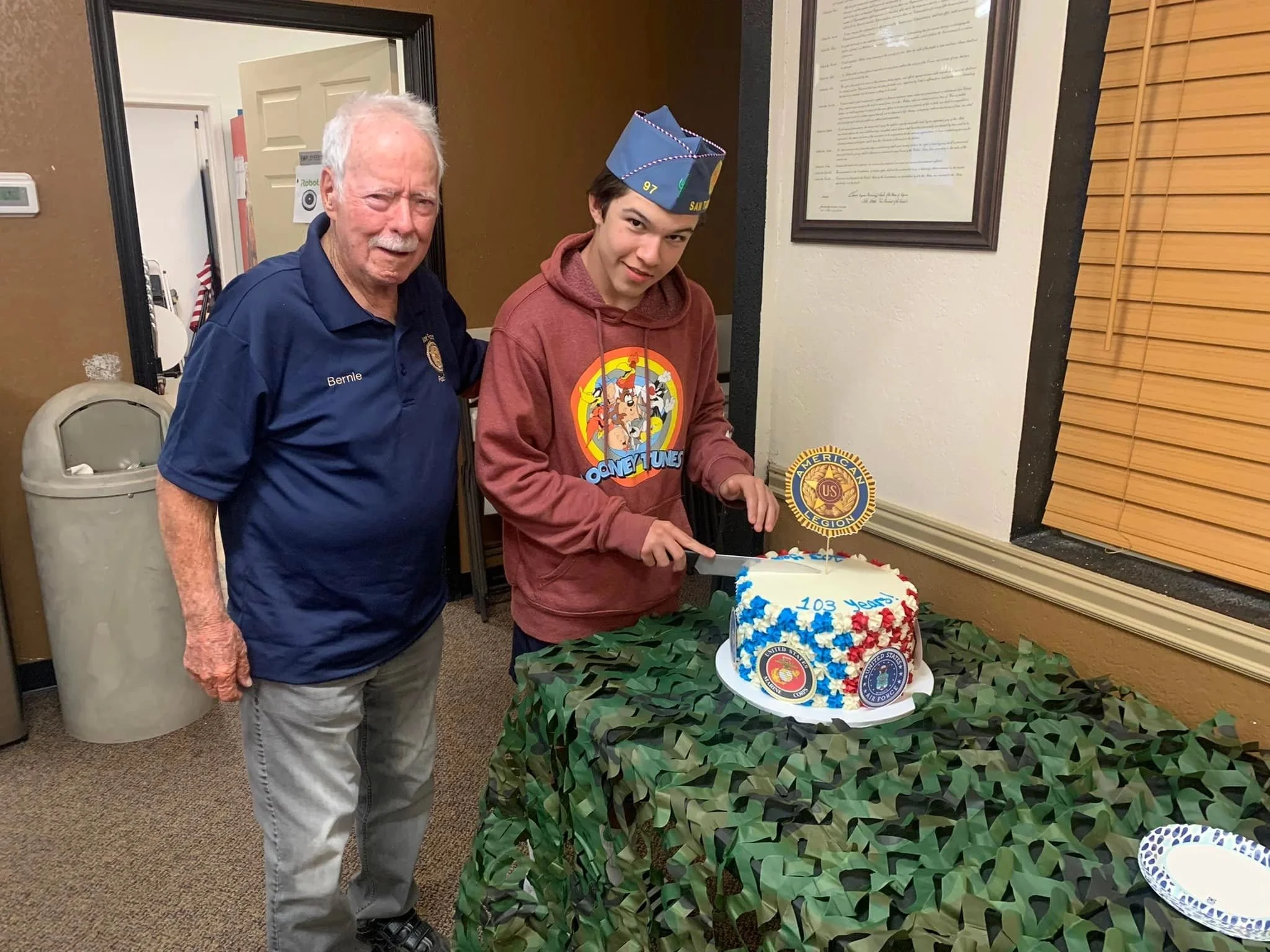 A birthday celebration with two men standing next to a cake decorated with American Legion symbols. One man is older with white hair, wearing a navy blue shirt with a name tag that reads "Bernie." The other younger man is wearing a red hoodie with a 