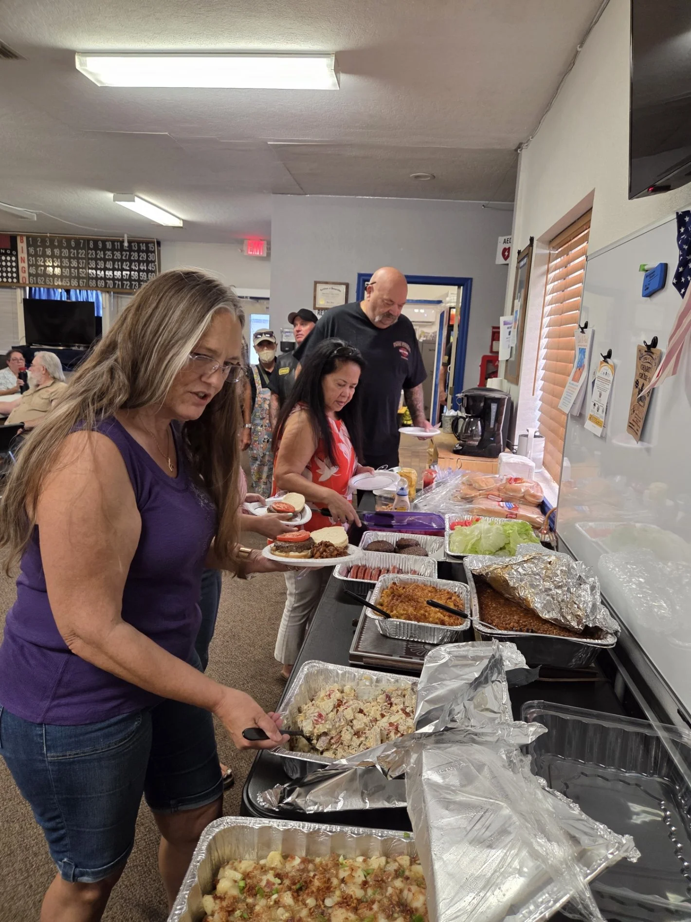 People lining up at a buffet table with various dishes including potato salad, pasta, and baked items in a community hall.