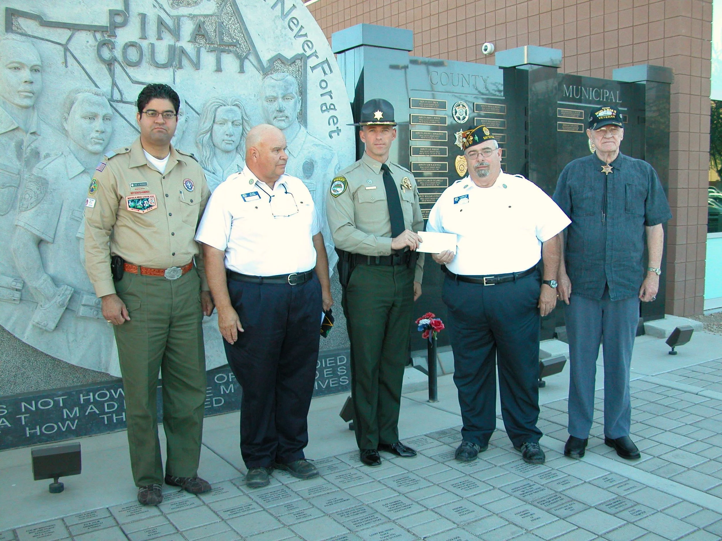 Six men standing in front of a memorial with engraved portraits and plaques. One man is dressed as a police officer, another in firefighter gear, and others in casual or uniform shirts. The memorial features a relief of faces and a large engraved map
