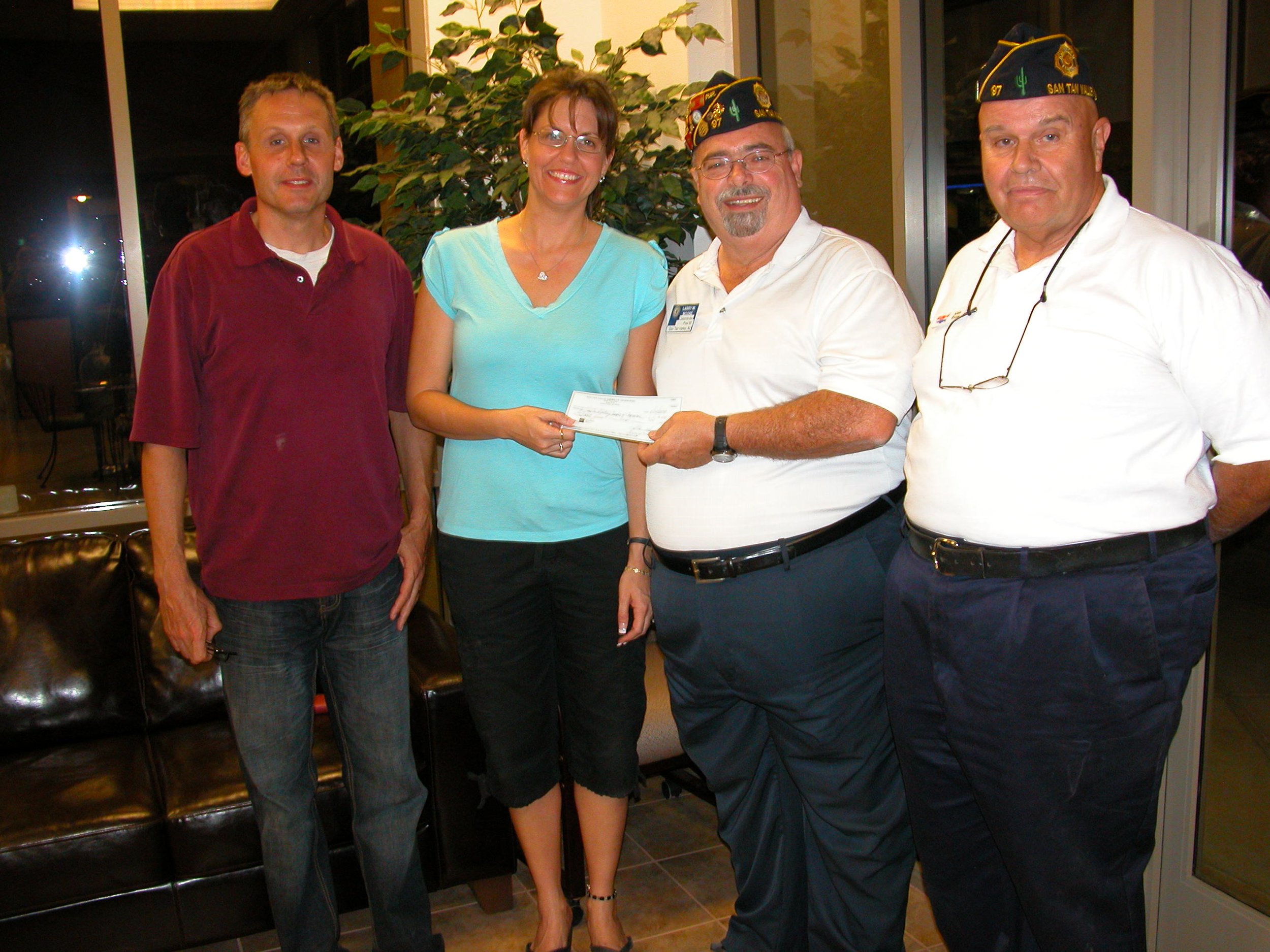 Four people standing side by side indoors at night. The woman in the center is holding a check and is flanked by two men wearing veteran caps with the words "San Tam Vets."