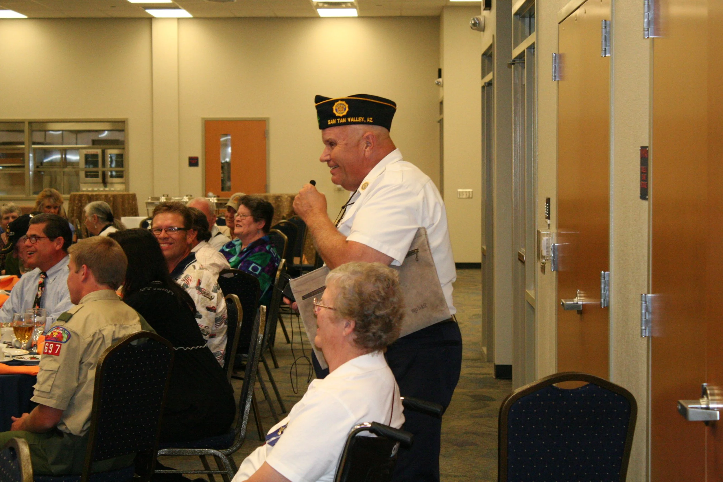 A man in a white uniform and a black hat with yellow trim and a badge, standing and speaking into a microphone at a banquet or formal gathering. The hat has the words 'San Tan Valley, AZ' embroidered on it. He is surrounded by seated guests, many of 