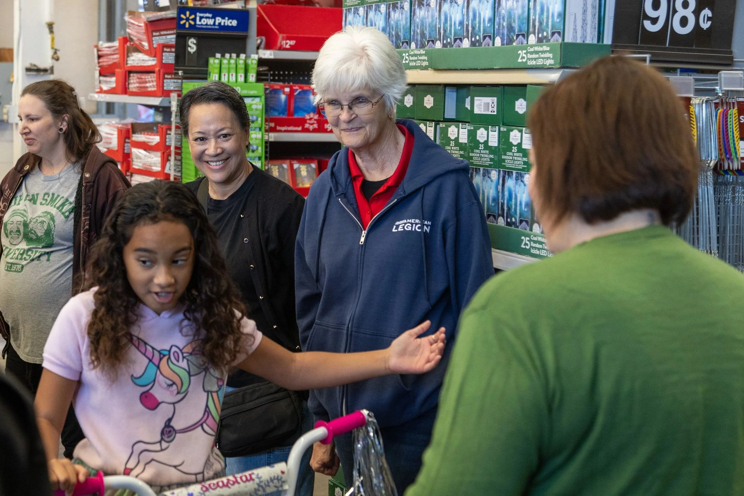 A group of people, including a young girl and a woman, are shopping inside a store. They are standing together, smiling and talking. The background shows shelves stocked with Christmas lights and other products.