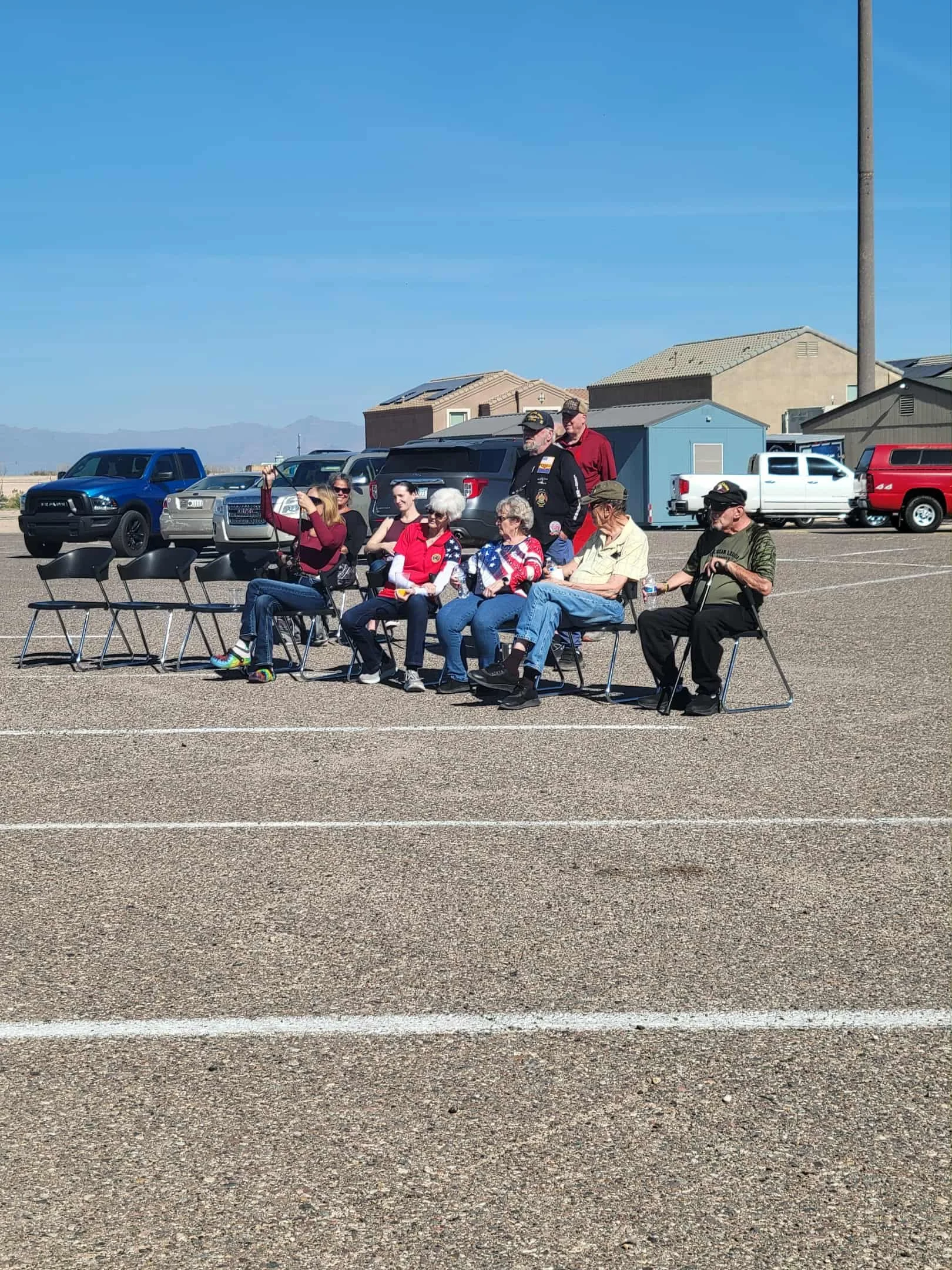 A group of people sitting and standing in a parking lot on a sunny day, with parked cars and houses in the background.