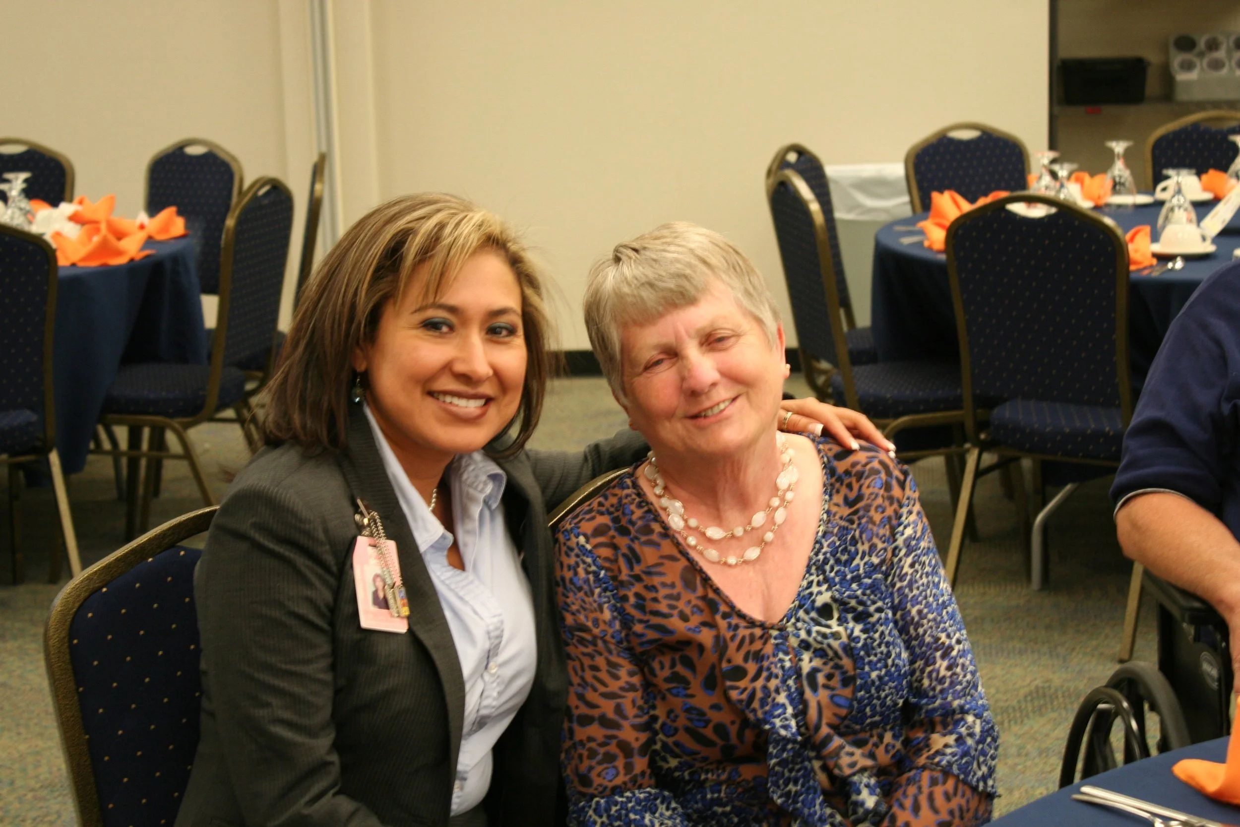 Two women sitting together at a banquet table, smiling at the camera. The woman on the left has short brown hair and is wearing a suit with a name badge. The woman on the right has short gray hair and is wearing a patterned top with a pearl necklace.