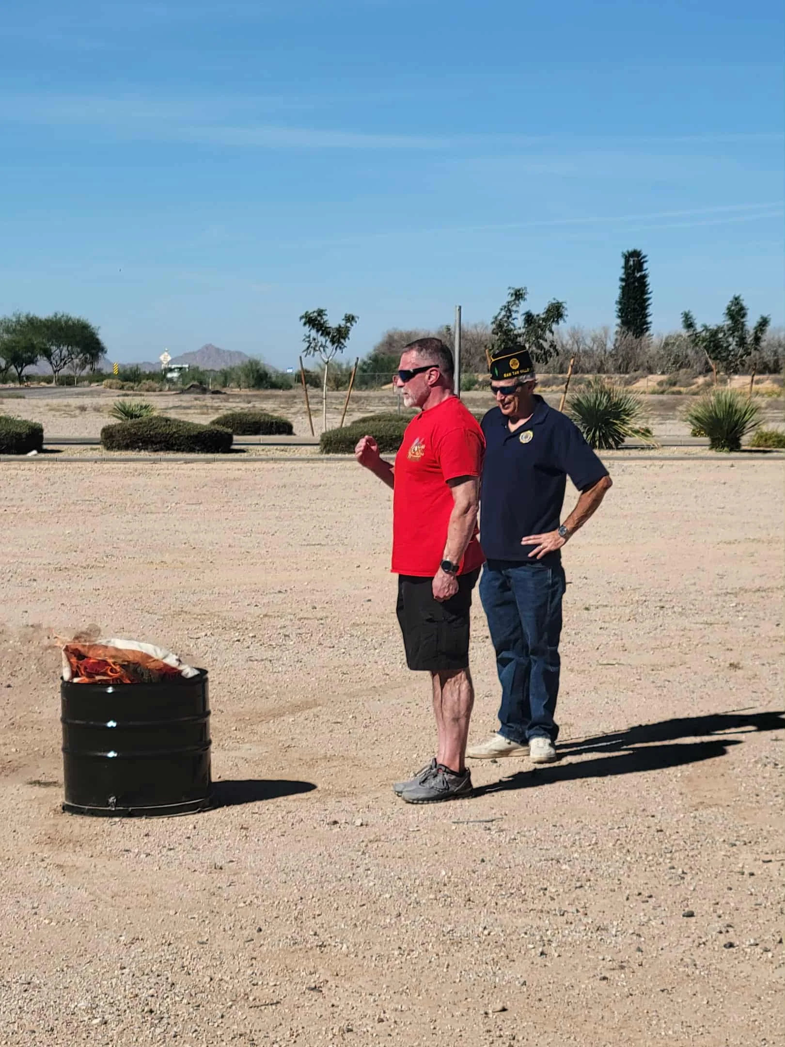 Two men standing outdoors near a barrel with a fire inside. One man is wearing a red shirt, shorts, sunglasses, and walking shoes. The other man is dressed in a blue polo shirt and cap, with his hands on his hips. The background features desert lands