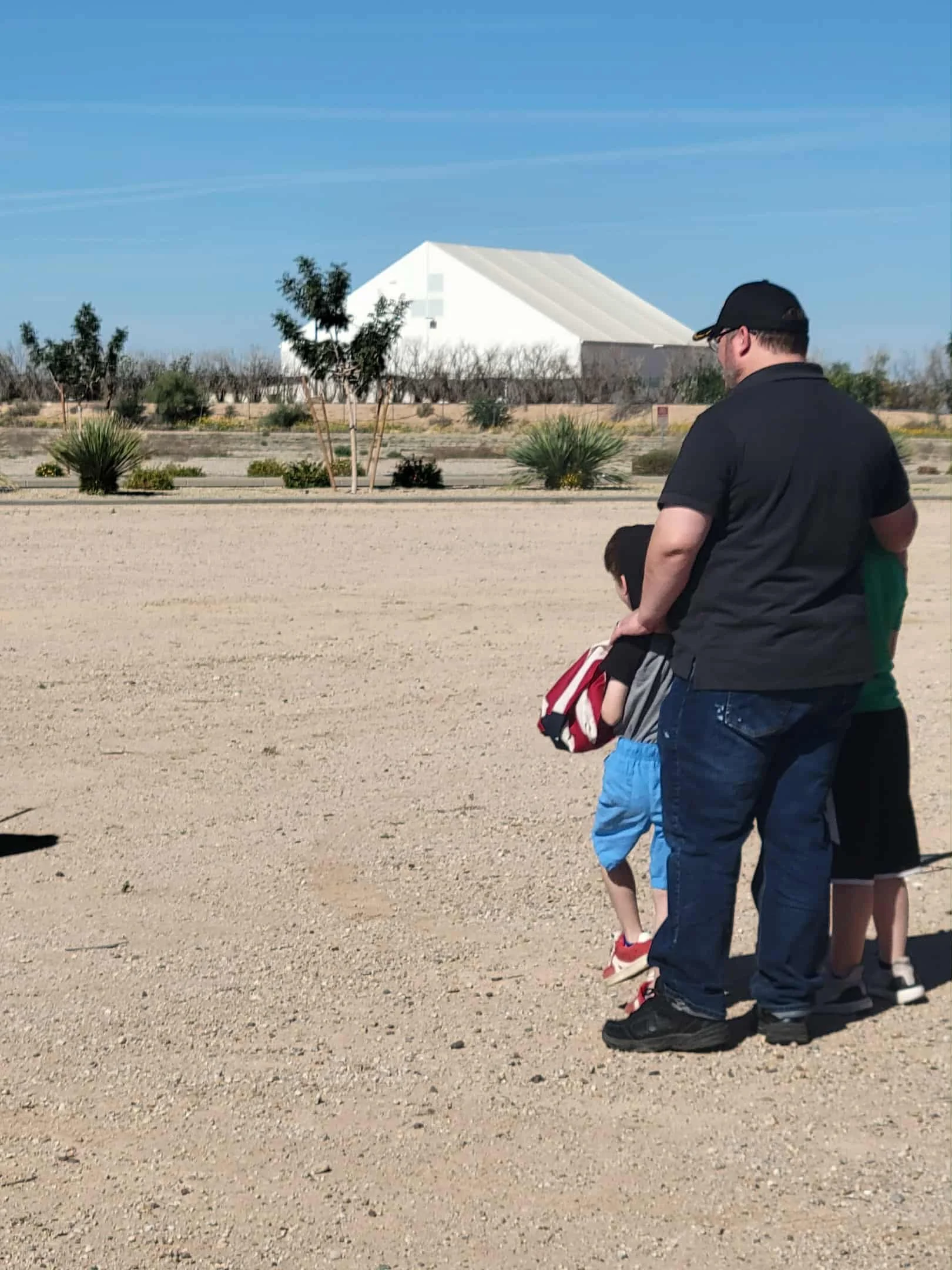 A man and children stand on a dirt path in a desert-like area, with desert plants and trees, and a large white building in the background under a clear blue sky.