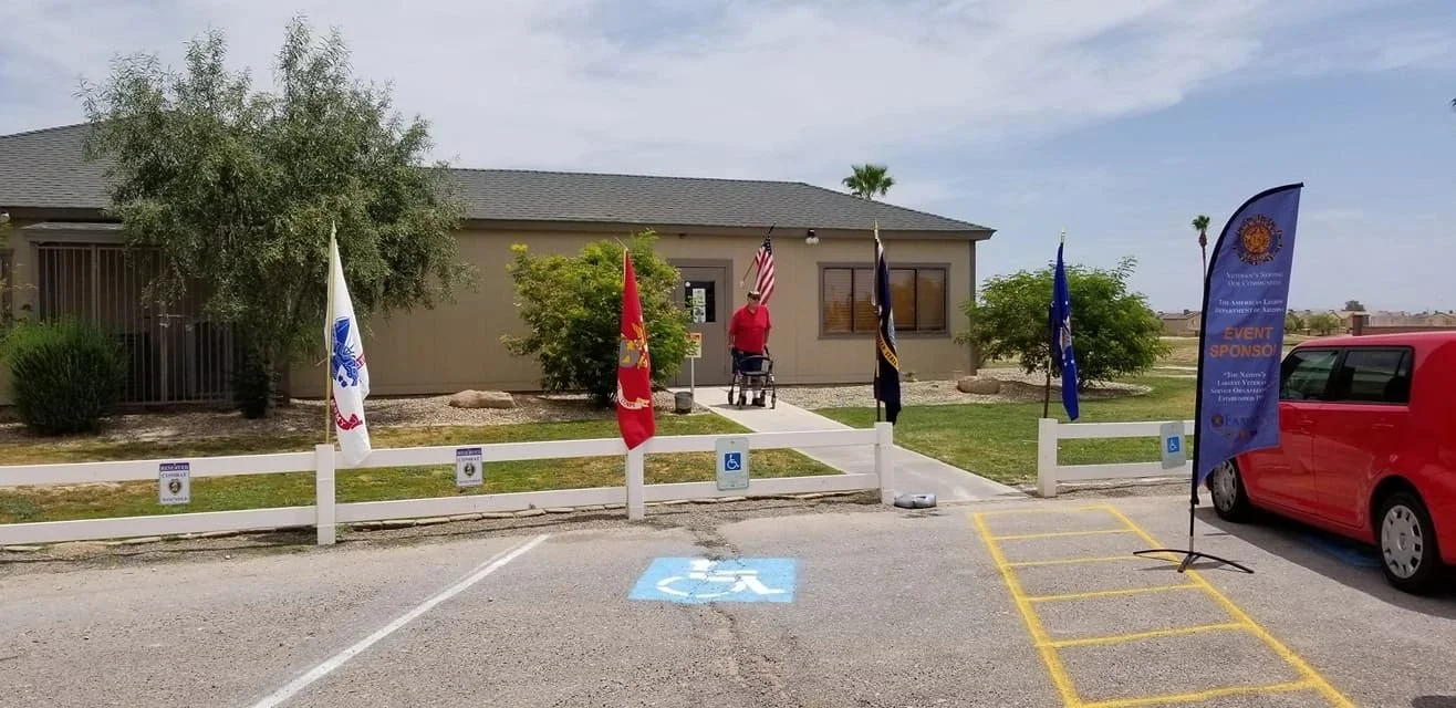 A small building or community center with flags, including American, military, and organizational flags, displayed outside. There is a person with a walker standing near the flags. The foreground shows a parking lot with a designated handicap parking