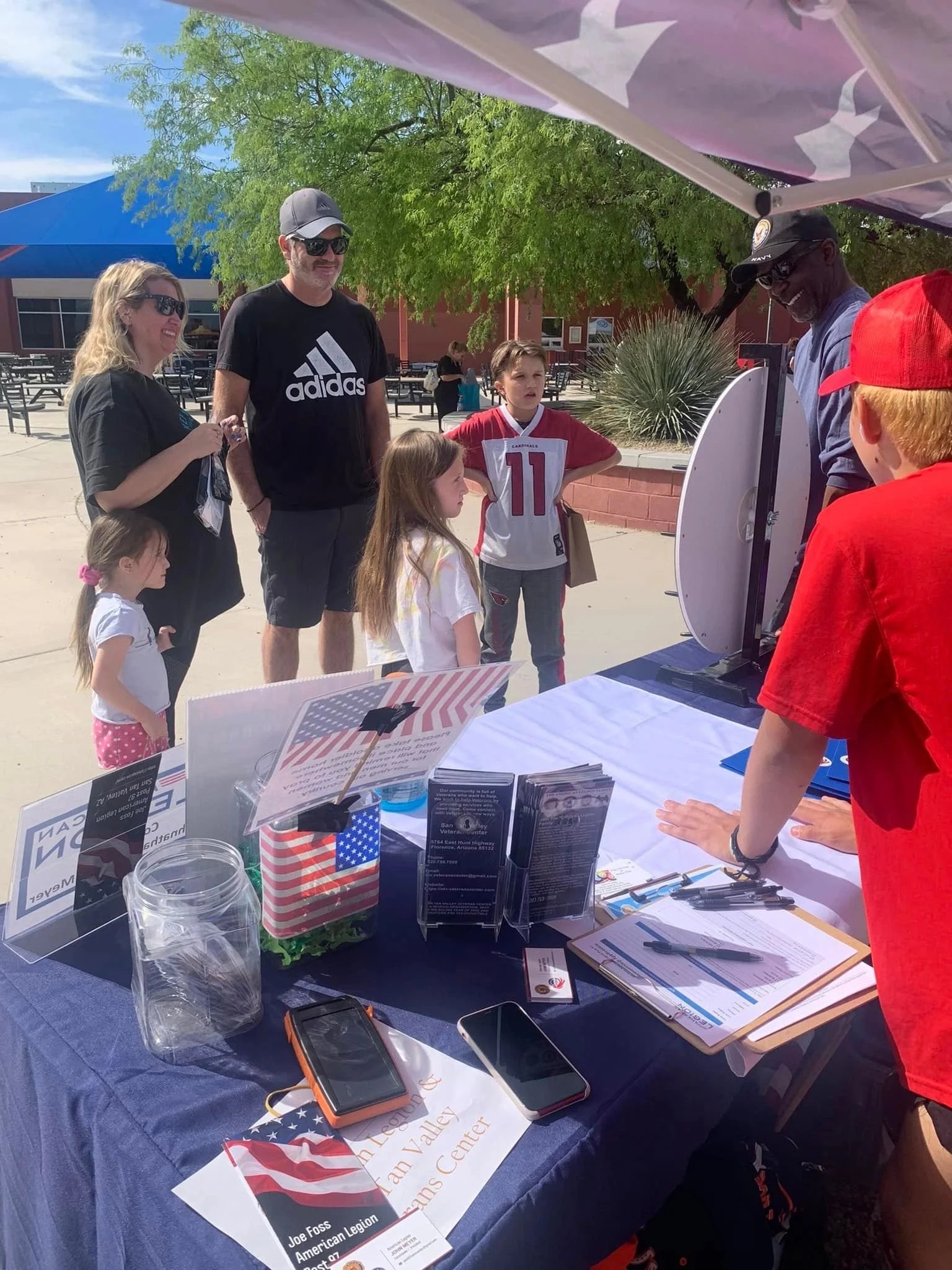 At an outdoor event, a table displays various informational materials, pamphlets, and a small American flag. Several people, including children and adults, are gathered around and engaging with a man behind the table, who appears to be explaining som