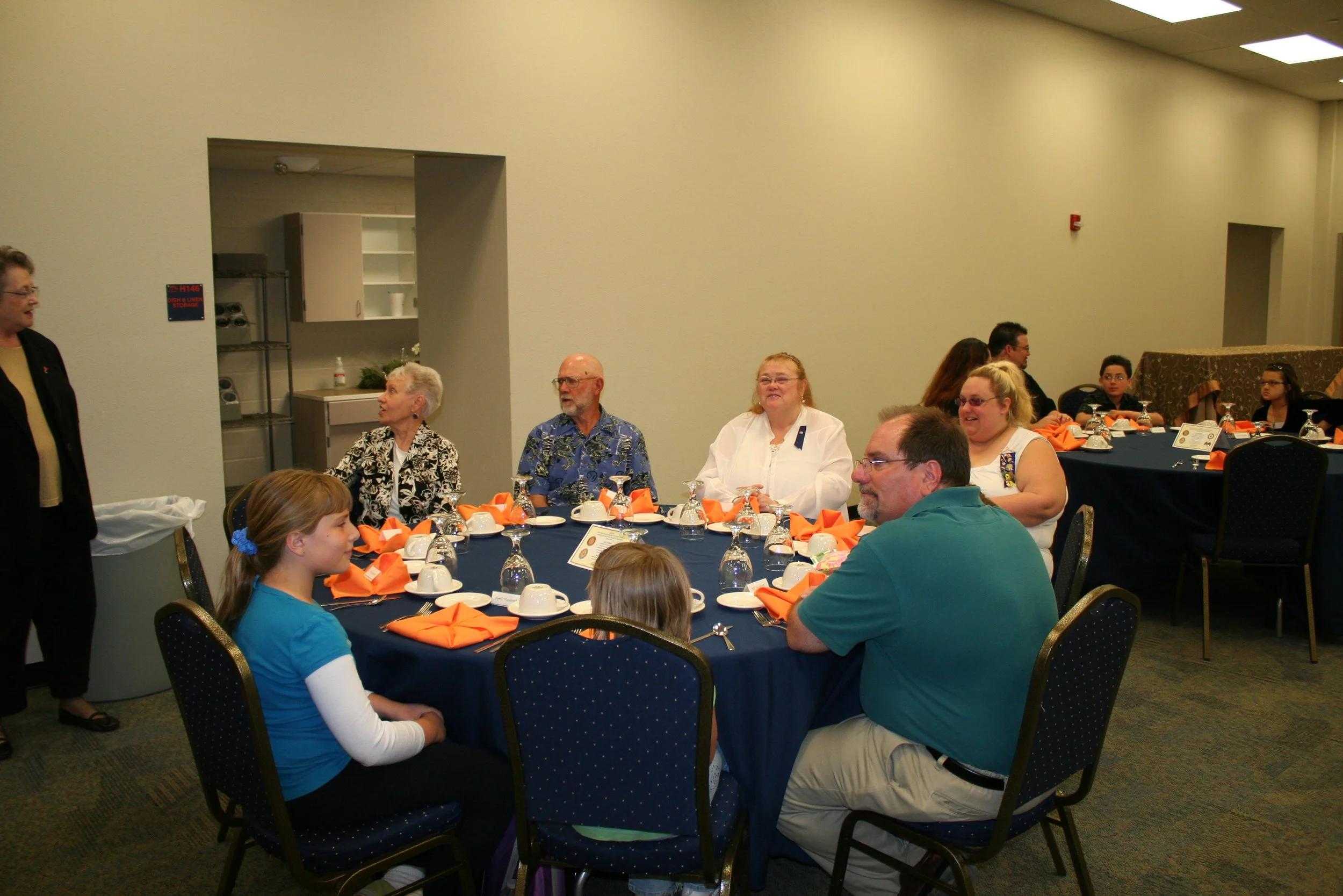 A group of diverse people sitting at a banquet table with orange napkins, white plates, cups, and silverware, attending an indoor event. Some people are smiling and talking, including a woman in a white blouse, a man in a Hawaiian shirt, and a young 