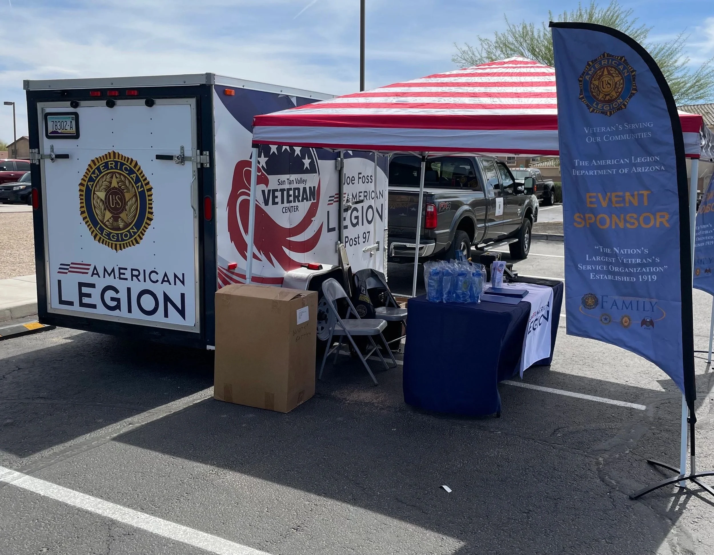 Outdoor setup for a Veteran's event with a trailer bearing the American Legion emblem, a booth with a red, white, and blue canopy, and a blue flag with American Legion insignia, in a parking lot with cars in the background.