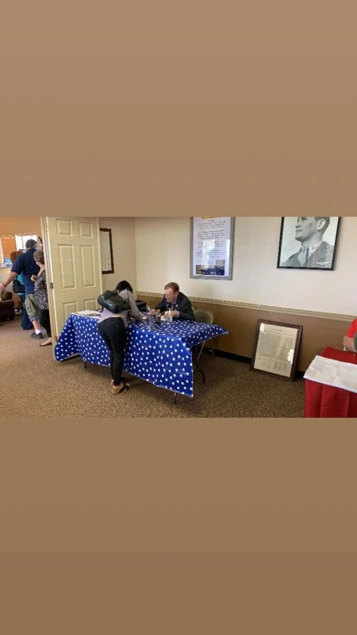 A man and woman sitting at a table with a blue star-spangled tablecloth, engaged in conversation. There are portraits on the wall behind them and a framed document leaning against the wall. A person with a black purse is standing at the table, and ot