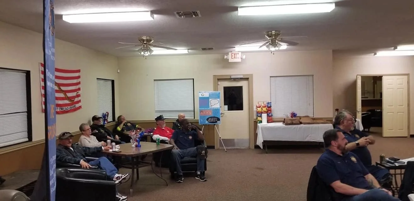 A group of mostly older men and women are seated and watching something in a room decorated with an American flag. There are tables with snacks and drinks, and posters on easels.