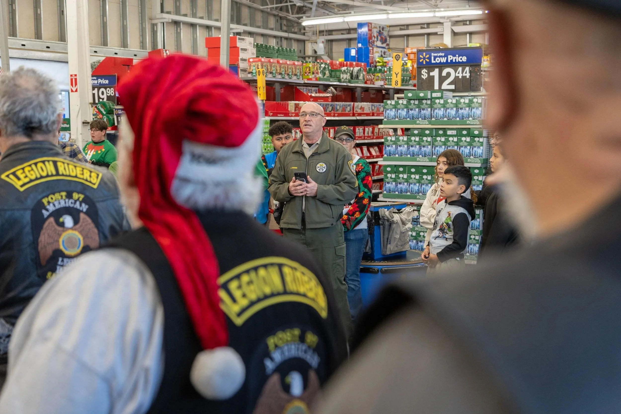 Group of people, including children and adults, gathered inside a retail store, listening to a man in a uniform speaking, with store shelves stocked with holiday-themed products in the background.