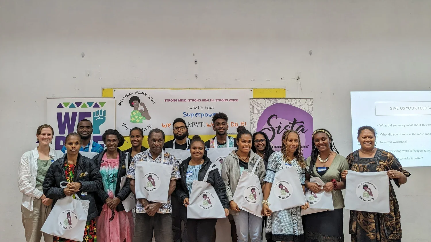 Group of workshop participants posing with “Melanesian Women Today” tote bags in front of women’s empowerment posters and presentation screen