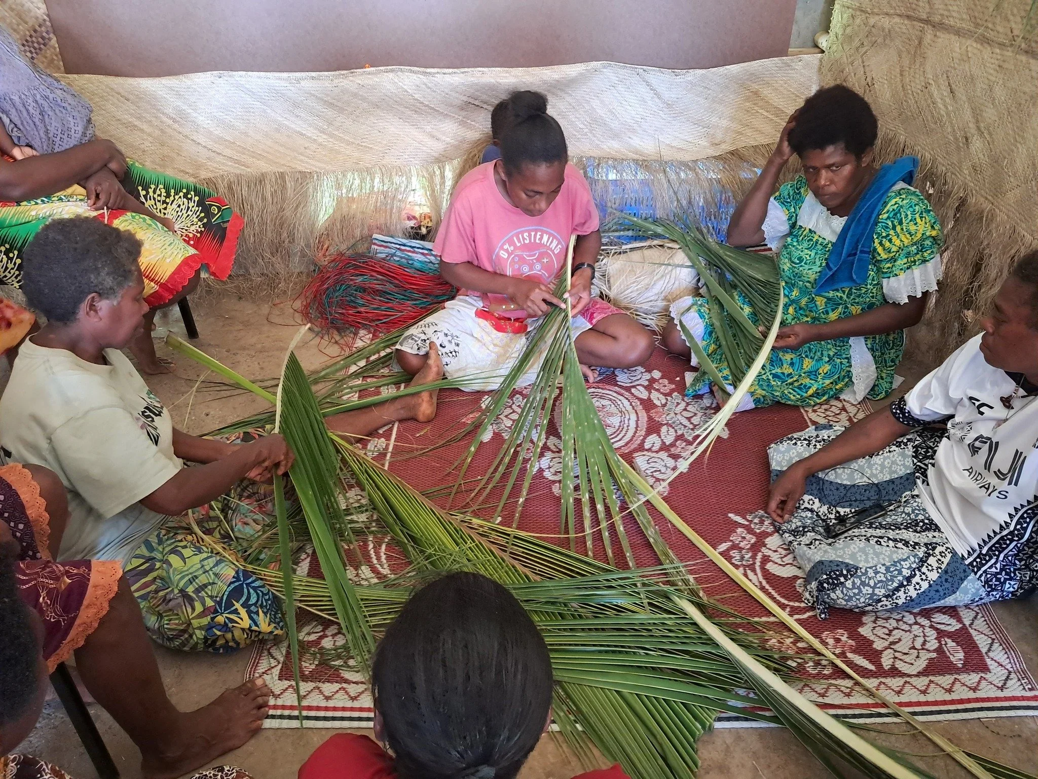 Group of melanesian women weaving palm leaves into traditional crafts inside a thatched shelter
