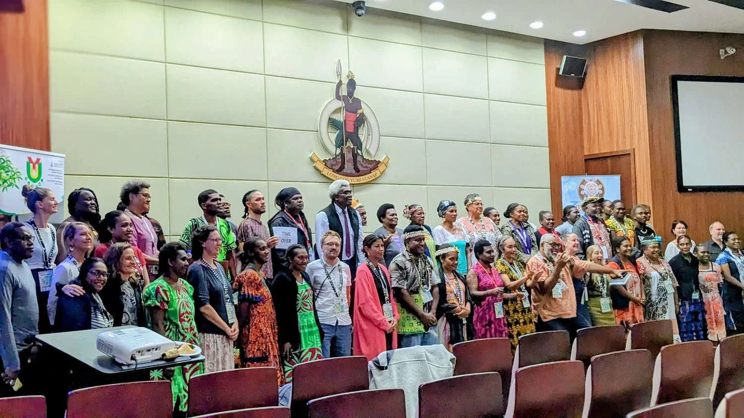 Large multicultural group posing in a university auditorium beneath the Papua New Guinea coat of arms