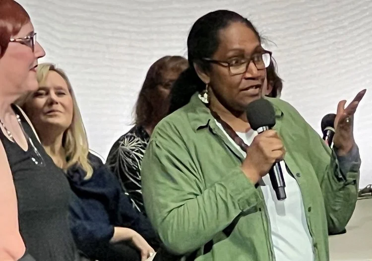 Mere Sovick in Odyssey Talent Show : Woman in a green shirt speaking into a microphone to an audience, with attendees standing behind her.
