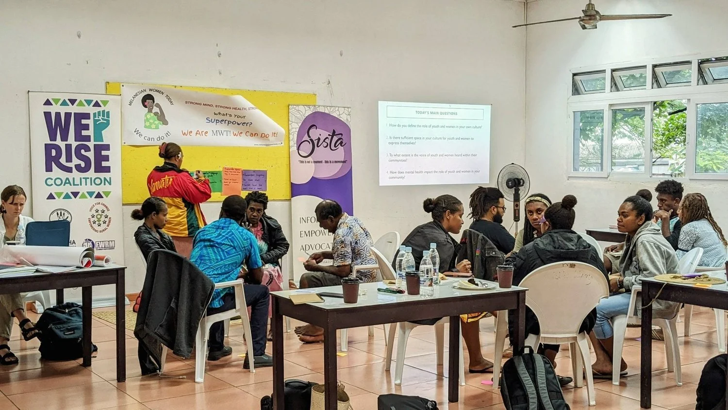 Youth workshop participants collaborate around tables in a community room decorated with We Rise Coalition and women’s empowerment posters.