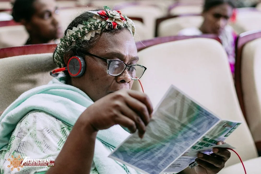 Woman wearing headphones and glasses reads a printed document in an auditorium.