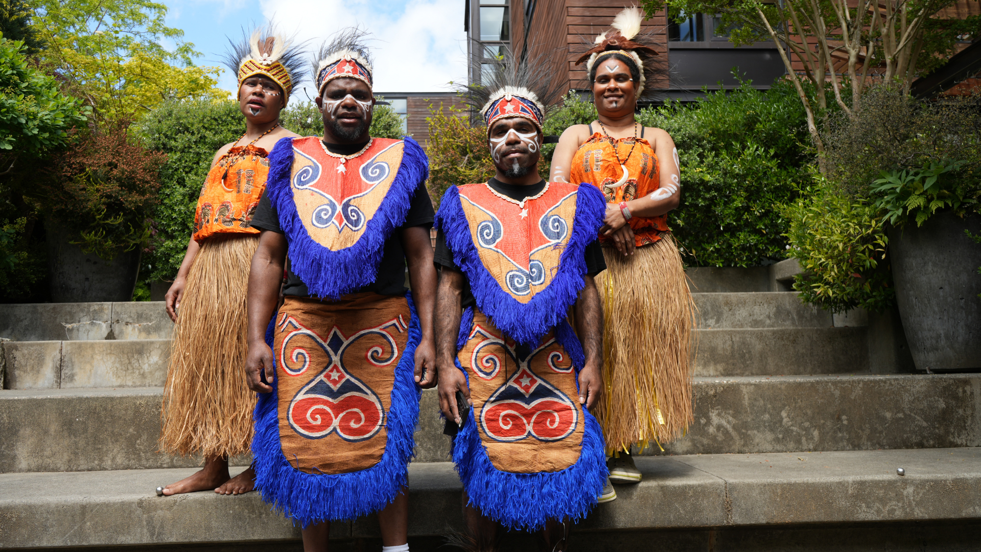 Four performers in traditional Papua New Guinean attire pose outdoors, wearing feathered headdresses, face paint, grass skirts, and colorful tasseled costumes.