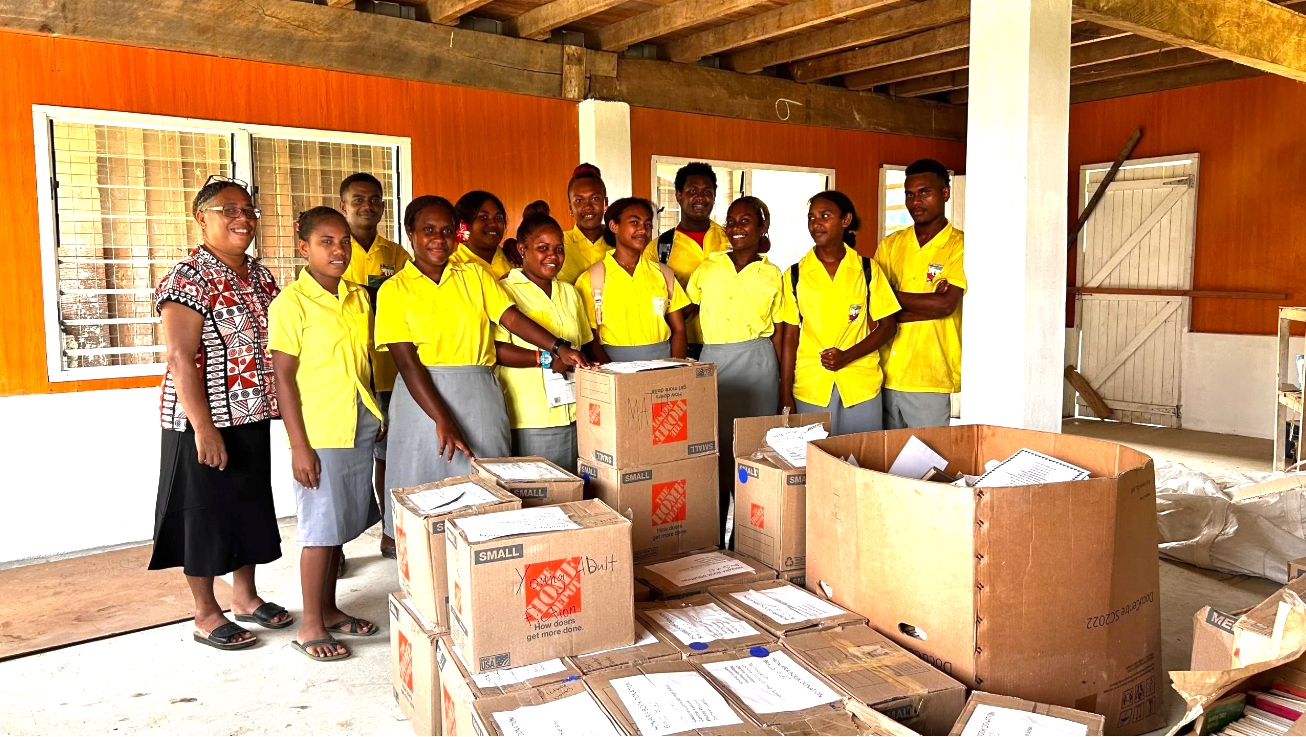 Solomon Islands Chapter : Group of students and a teacher in yellow uniforms standing behind boxes of donated supplies indoors
