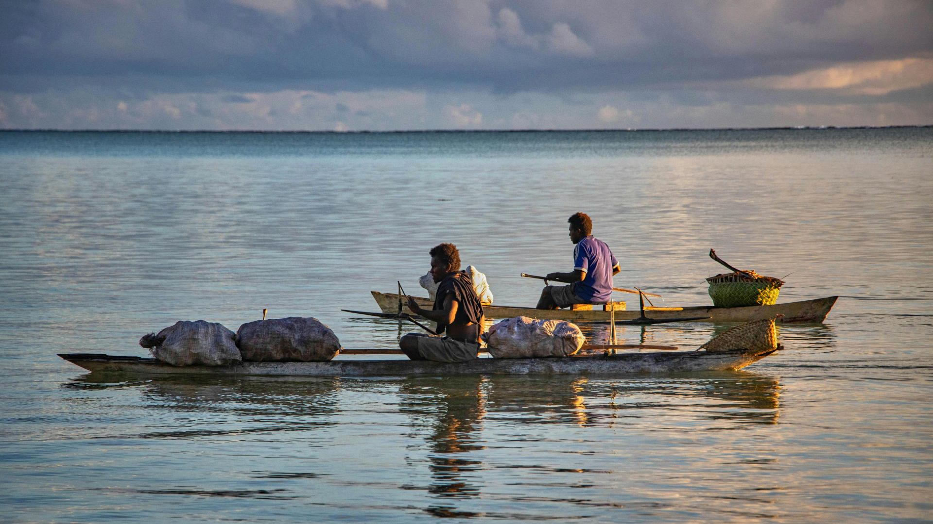 Two fishermen paddling narrow wooden boats loaded with sacks and woven baskets on calm melanesia water