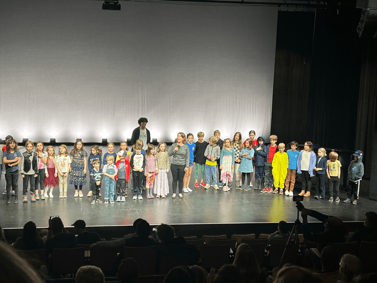 Odyssey Talent Show :  Children and teachers lined up on a theater stage during a school performance, with an audience seated in front.
