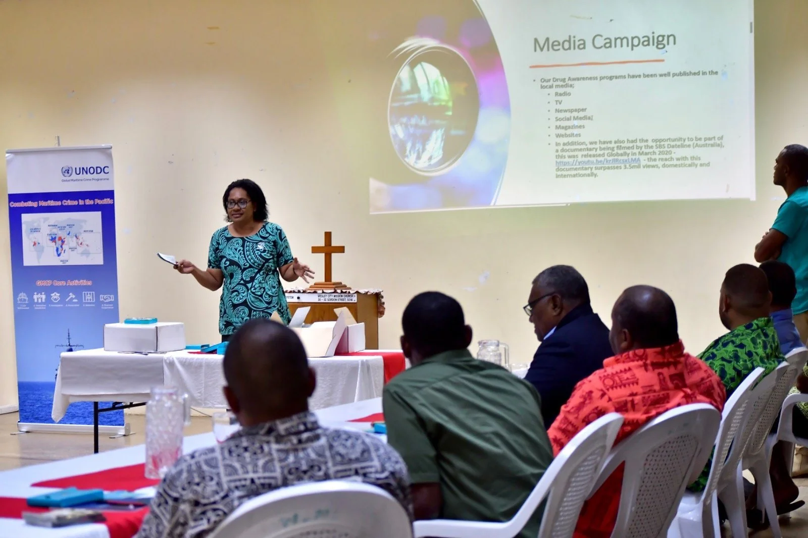 Woman presenting a media campaign slide at a UNODC drug-awareness meeting with seated attendees