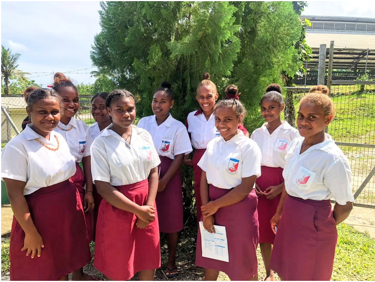 Solomon Islands Chapter : Group of nine smiling schoolgirls in white shirts and burgundy skirts posing outdoors.
