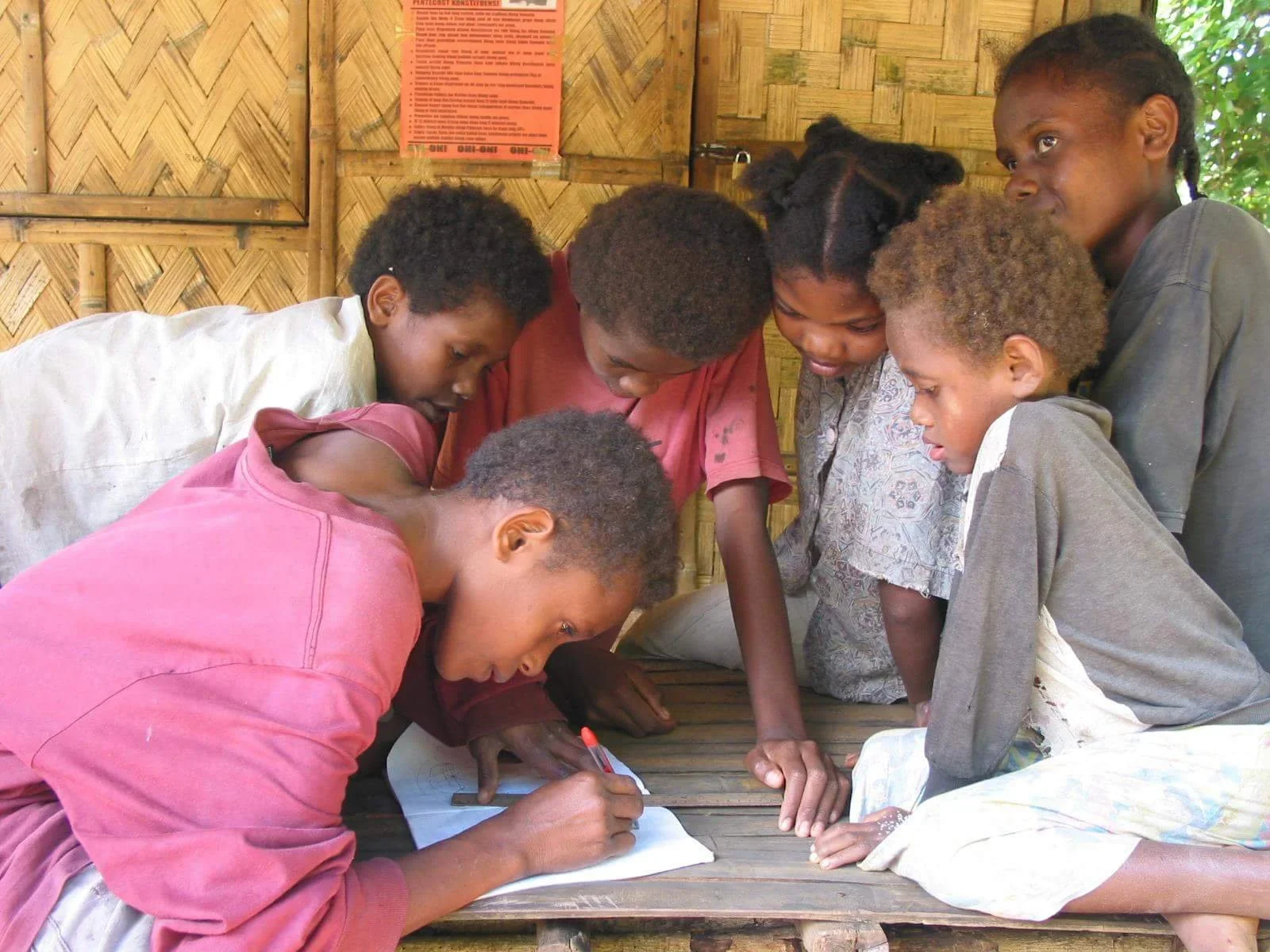 Vanuatu Children gathered around a student writing in a notebook inside a bamboo-walled classroom.