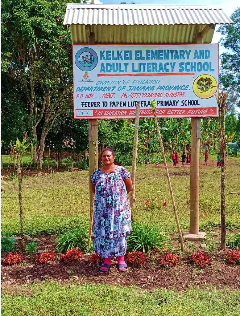 Woman standing beneath a sign for Kelkei Elementary and Adult Literacy School in Papua New Guinea.