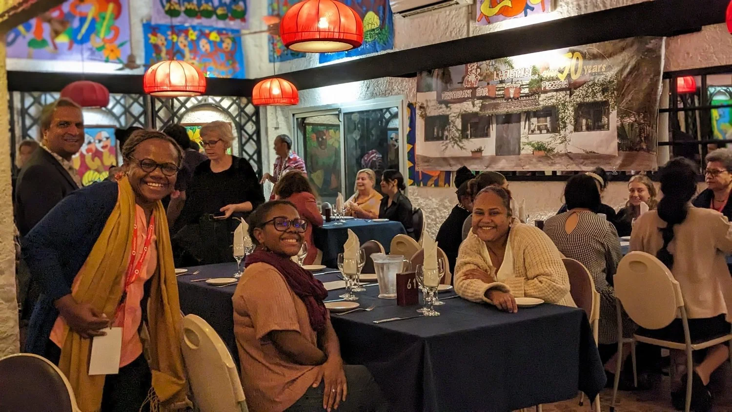 Guests dining and posing for a group photo inside a colorful restaurant with red pendant lights and artwork.