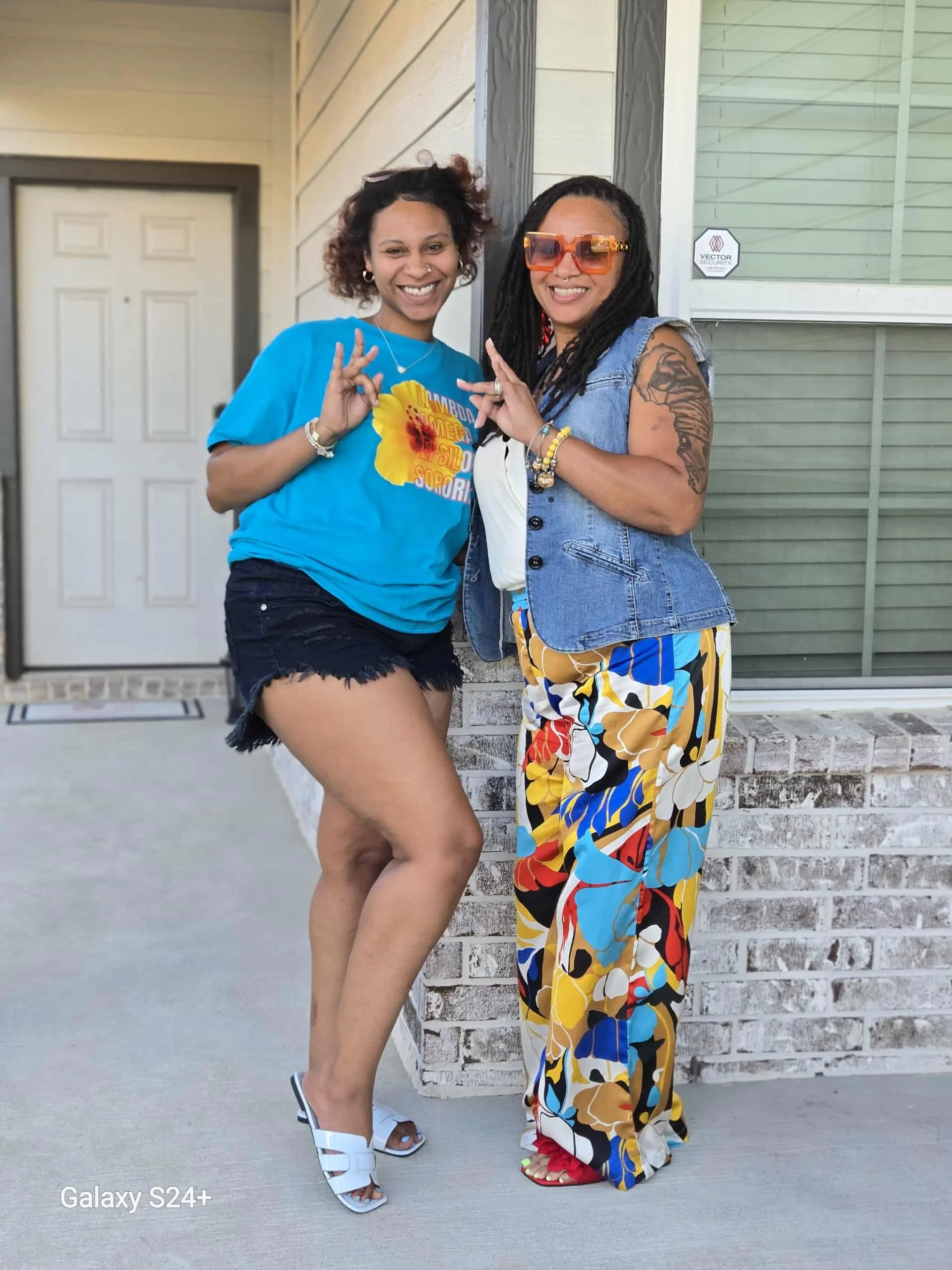 Two women smiling and posing side by side in front of a house. One woman has curly hair, wears a blue t-shirt with a floral design and black shorts, and white sneakers. The other woman has long dreadlocks, large orange sunglasses, tattoos on her arm,