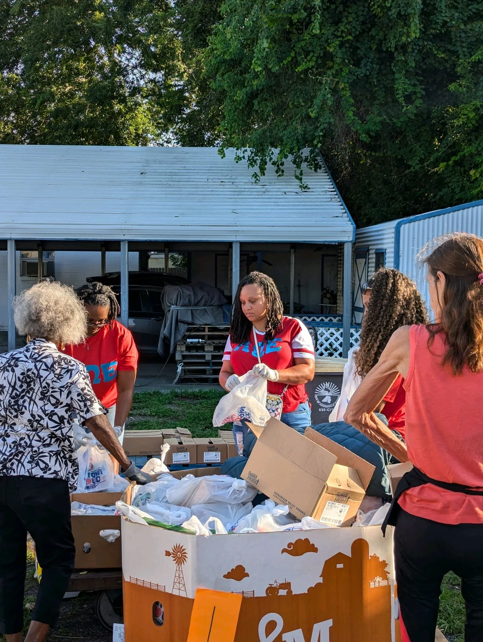 A group of people gather around a table with boxes and bags of food, possibly at a community food distribution event in a backyard with a house and trees in the background.