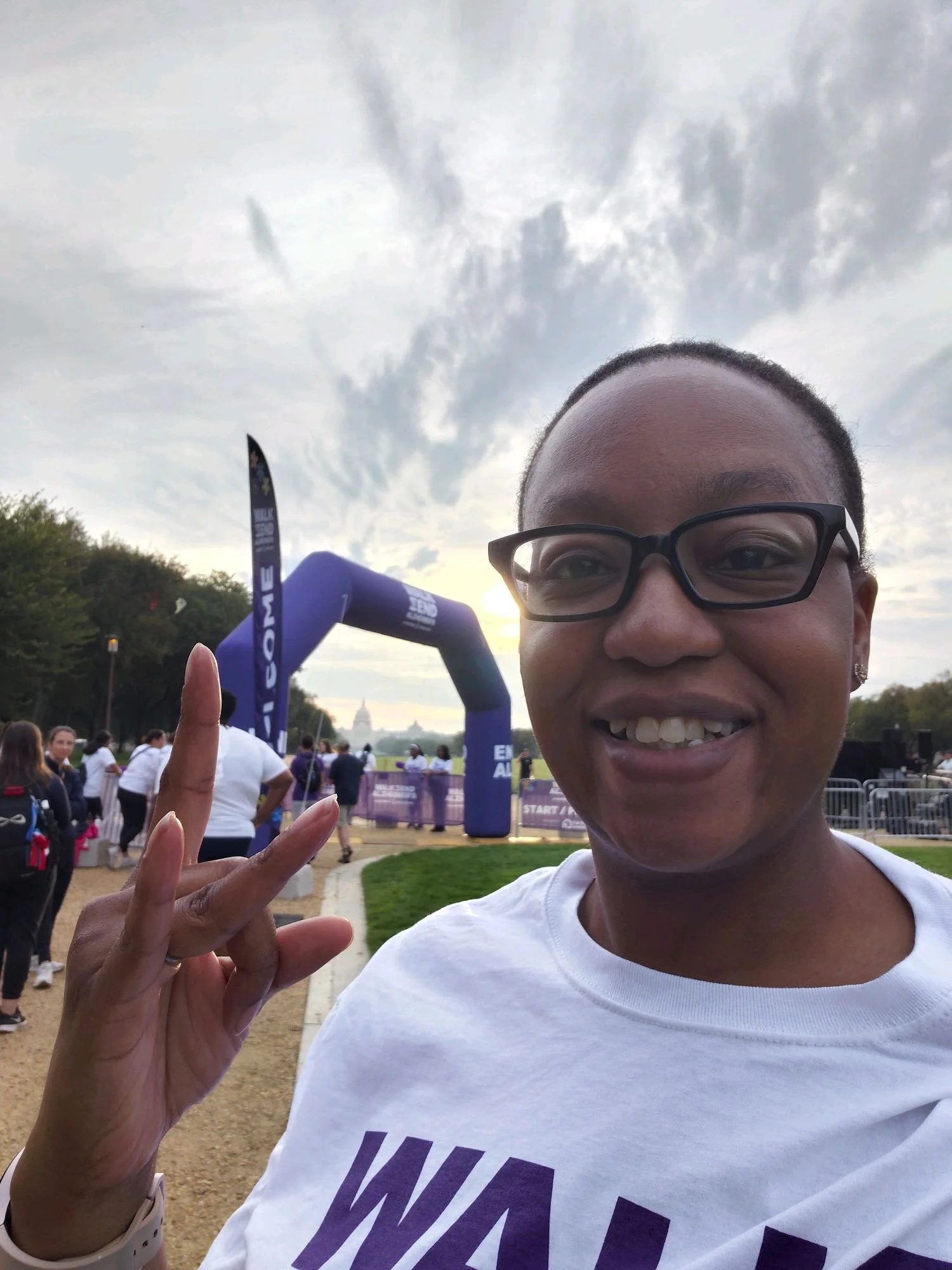 Person smiling and holding up a peace sign at a race event with a crowd and finish line arch in the background.