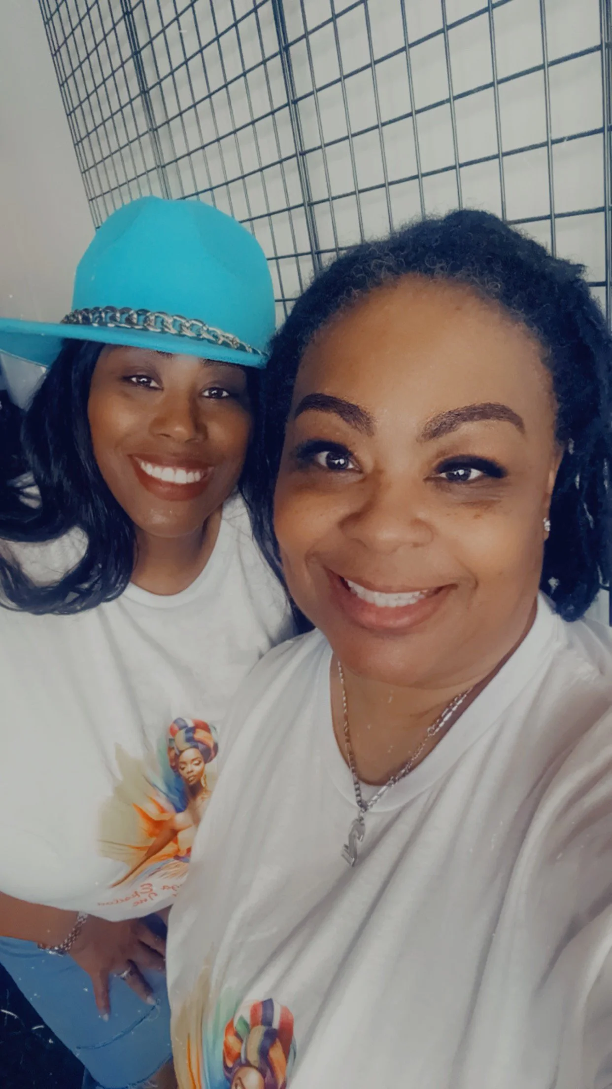 Two women smiling, one wearing a light blue hat with a silver chain and a white t-shirt, and the other with short curly hair, wearing a white t-shirt and a necklace, standing in front of a wire grid background.