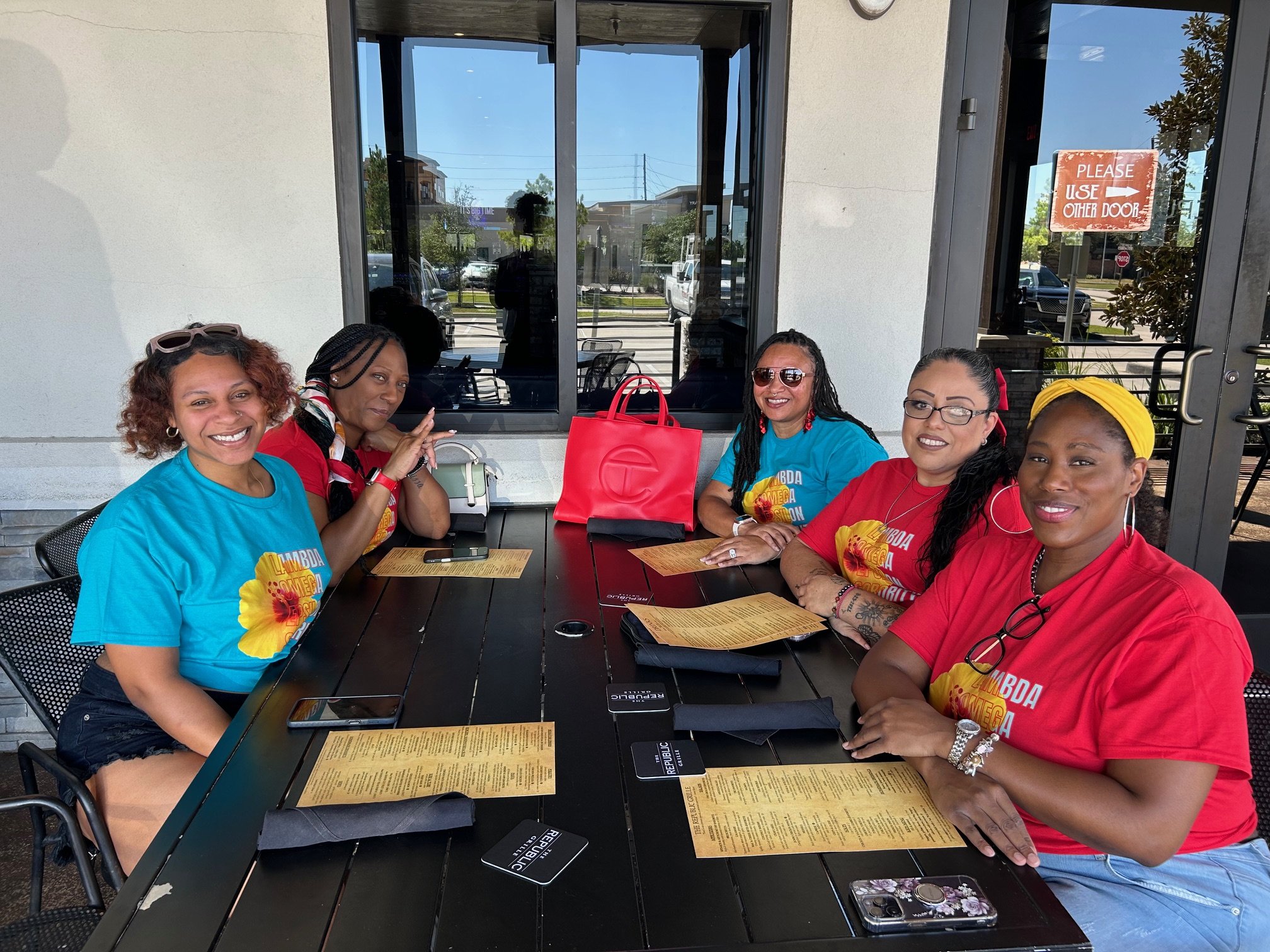 Group of six women sitting around a table outside of a restaurant or cafe, smiling and enjoying each other's company. The women are wearing colorful tops and sunglasses, and there are menus, phones, and a red handbag on the table. The background features a large window, an outdoor seating area, and a sign on the door that says 'Please Use Other Door'.